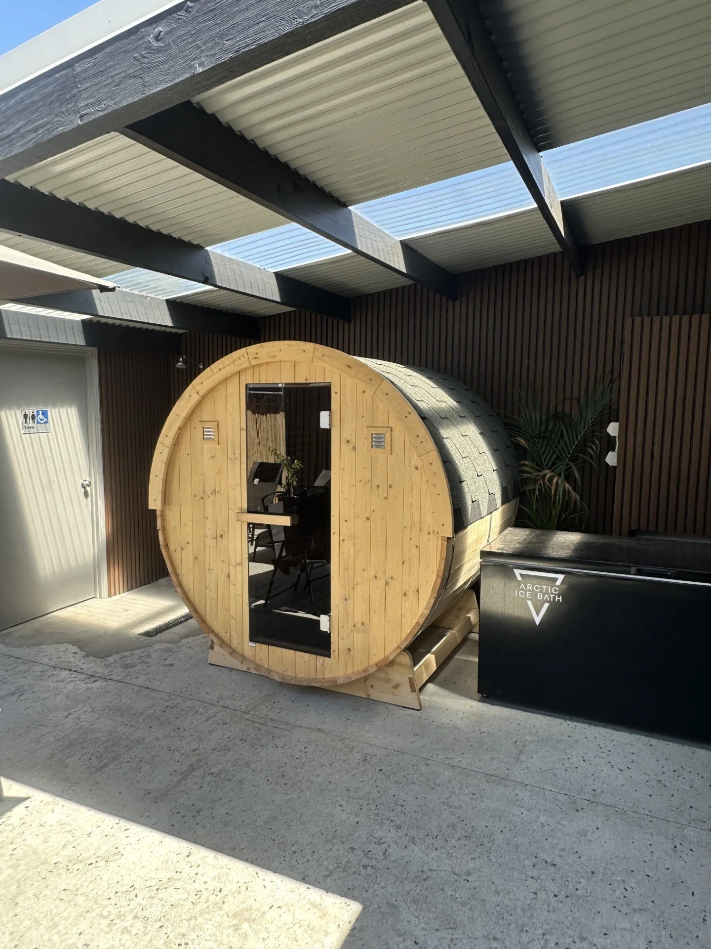 A wooden barrel-shaped sauna with a small window and black door, placed outdoors on a paved area, next to a black trash bin labeled 'Arctic Ice Bath'. There is a tree with leaves in the background, and a dark wooden wall with a corrugated metal roof overhead.