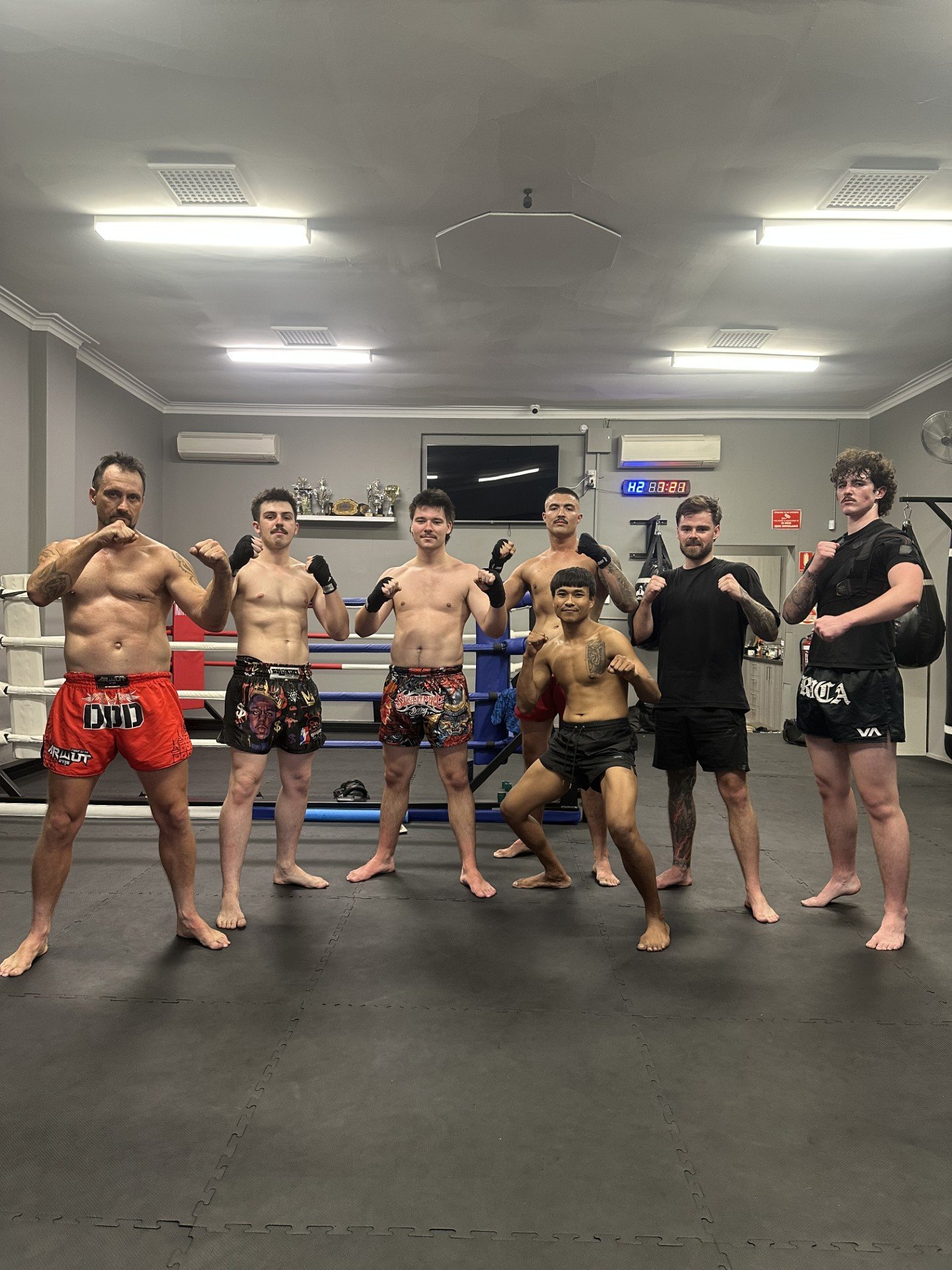 Group of nine people in a boxing gym, posing with fists up in fighting stance, standing and kneeling in front of boxing ring.