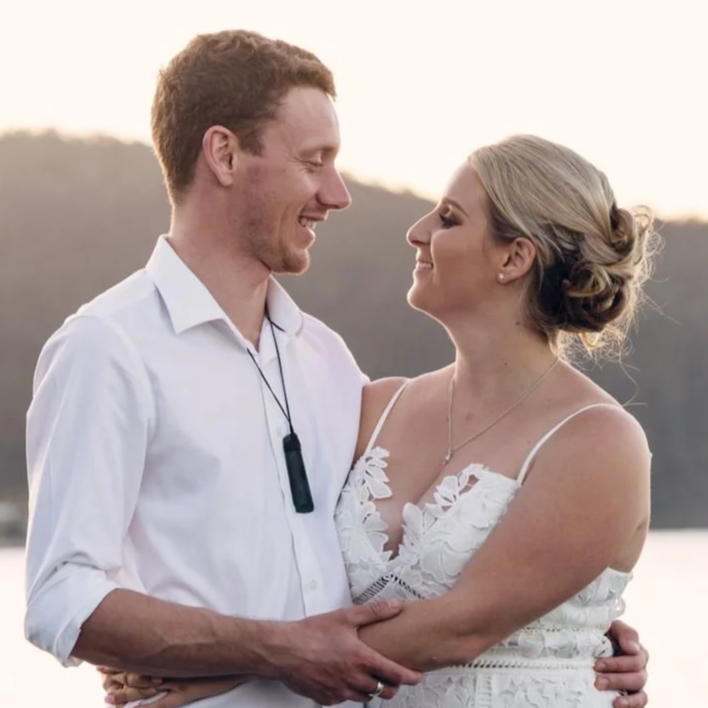 A couple stands close together outdoors during sunset, smiling and gazing at each other, with a lake and trees in the background. The man is wearing a white shirt and a black lanyard, and the woman is in a white dress with lace details and her hair styled in an updo.