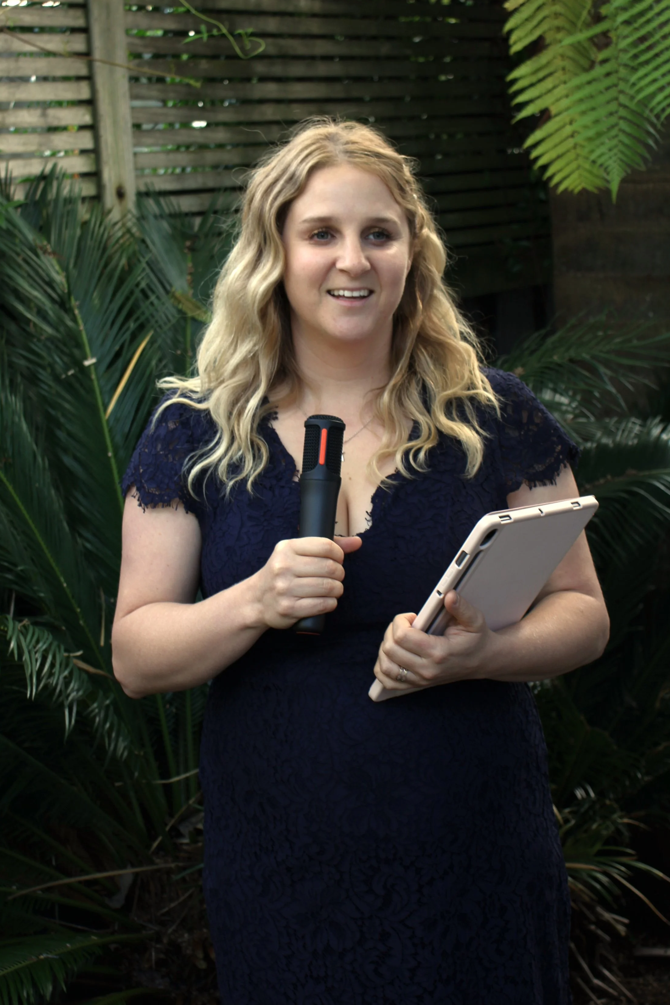 A woman (Nicola) with long blonde hair holding a microphone and a tablet, standing outdoors among green plants.