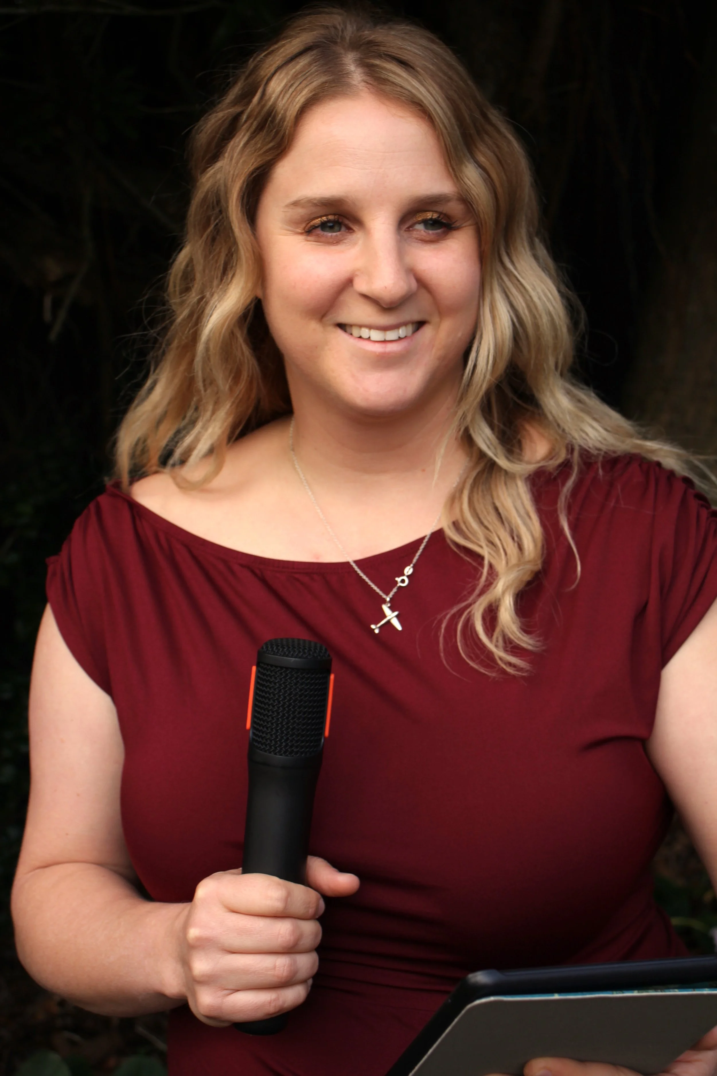 A woman (Nicola) with wavy blonde hair, wearing a red dress and a cross necklace, holding a black microphone in her right hand and a tablet in her left hand, standing outdoors against a dark background.