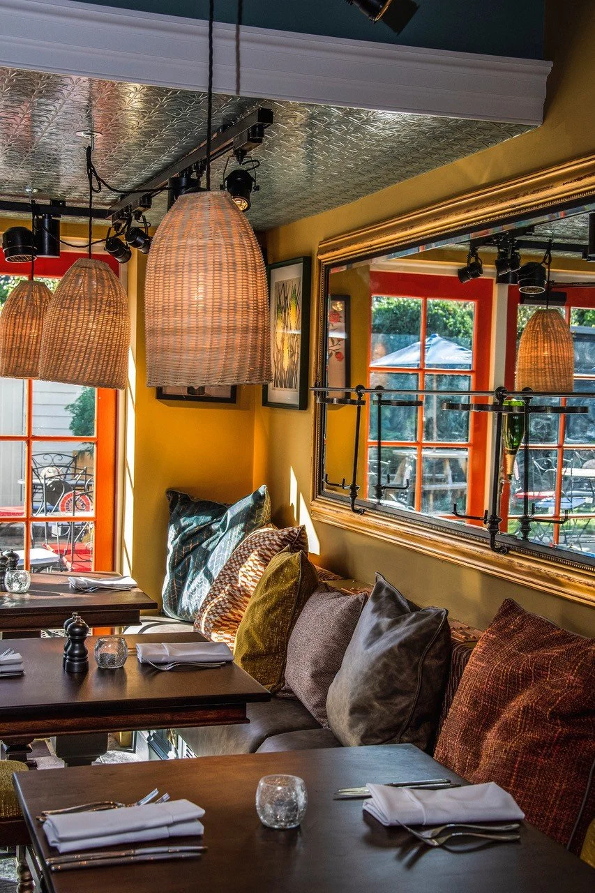 Interior of a cozy restaurant with tables set with white napkins, glass candle holders, and pepper shakers. The space features large windows with orange frames, hanging woven pendant lights, and colorful pillows on a bench along the wall. Outside, pa