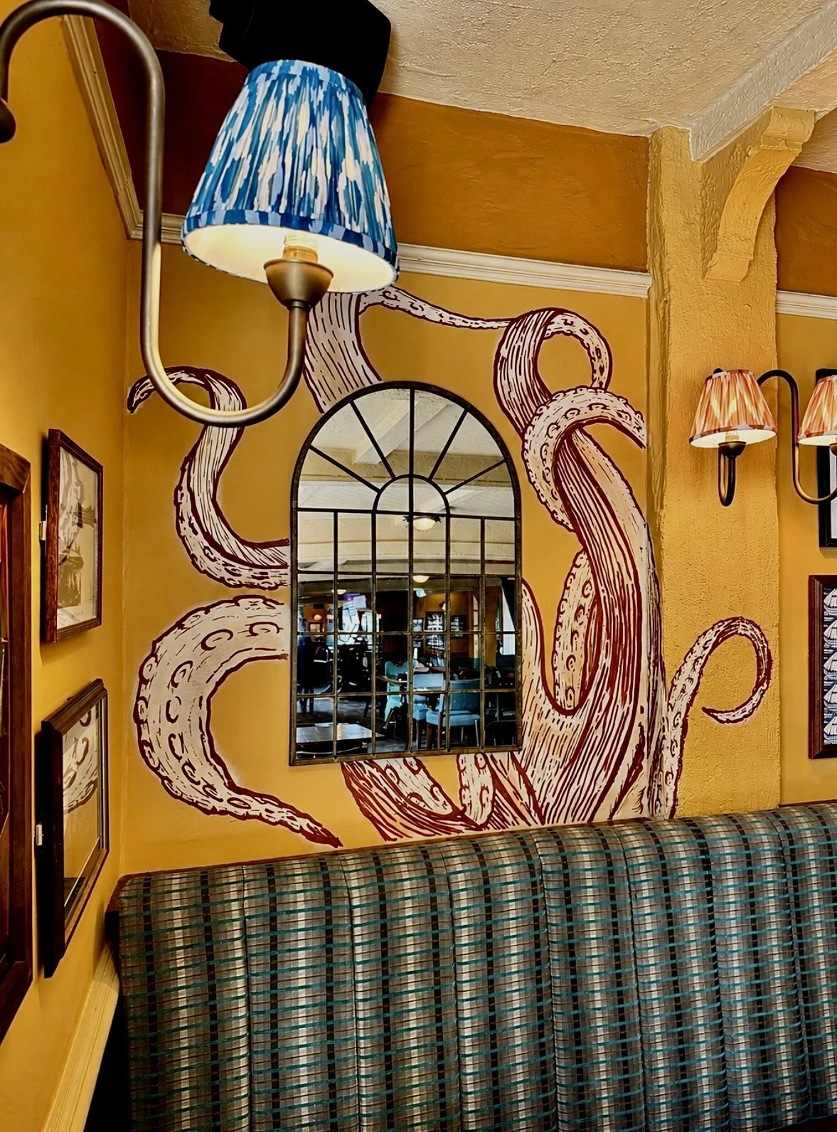 Decorative restaurant wall with a large mural of an octopus tentacle, a arched window mirror, and framed pictures, with colorful lamps and a plaid upholstered bench.