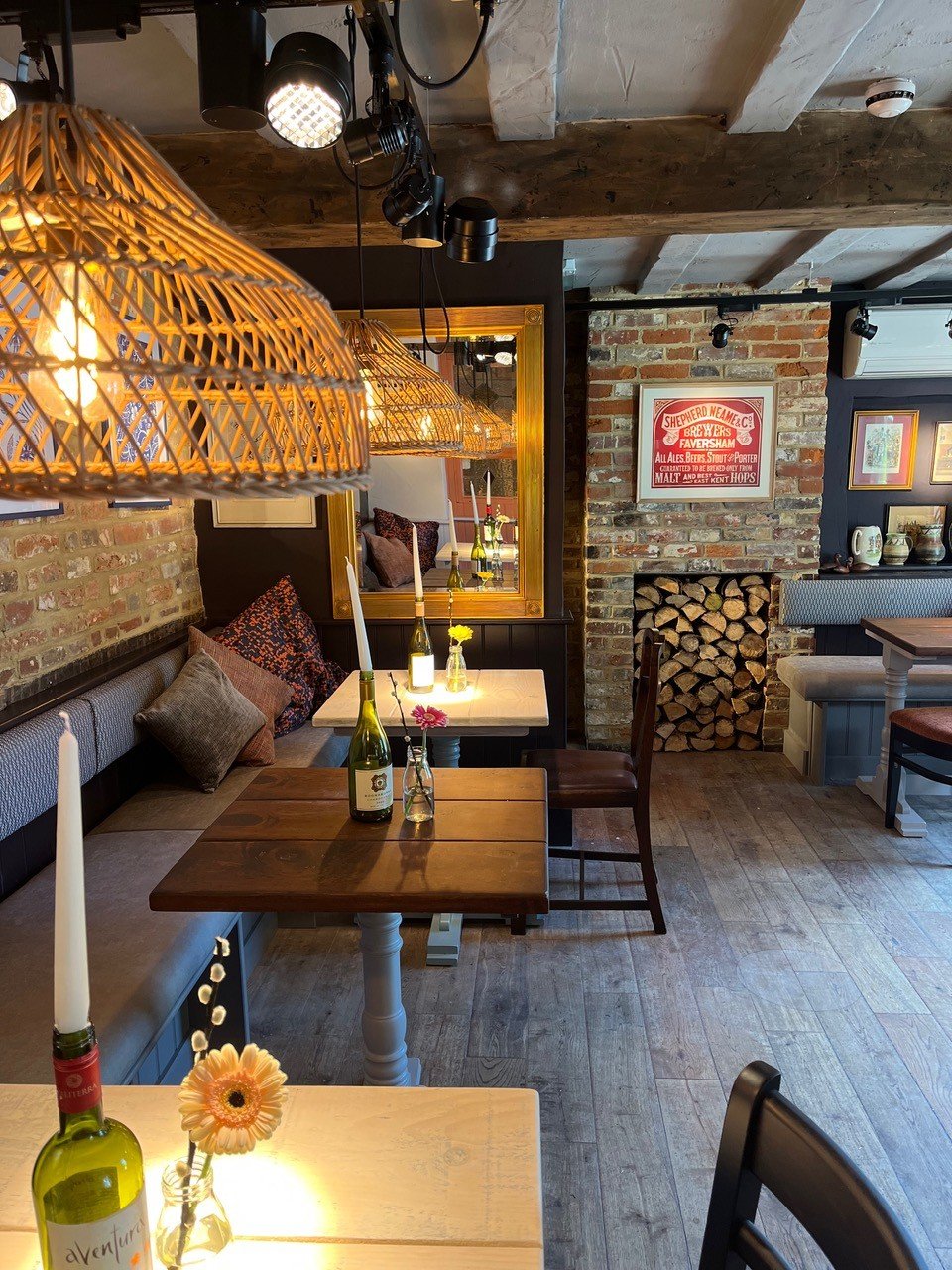 Cozy restaurant interior with brick walls, wooden beams, and vintage decor. The tables are decorated with candles and flowers, and wicker pendant lights hang from the ceiling.