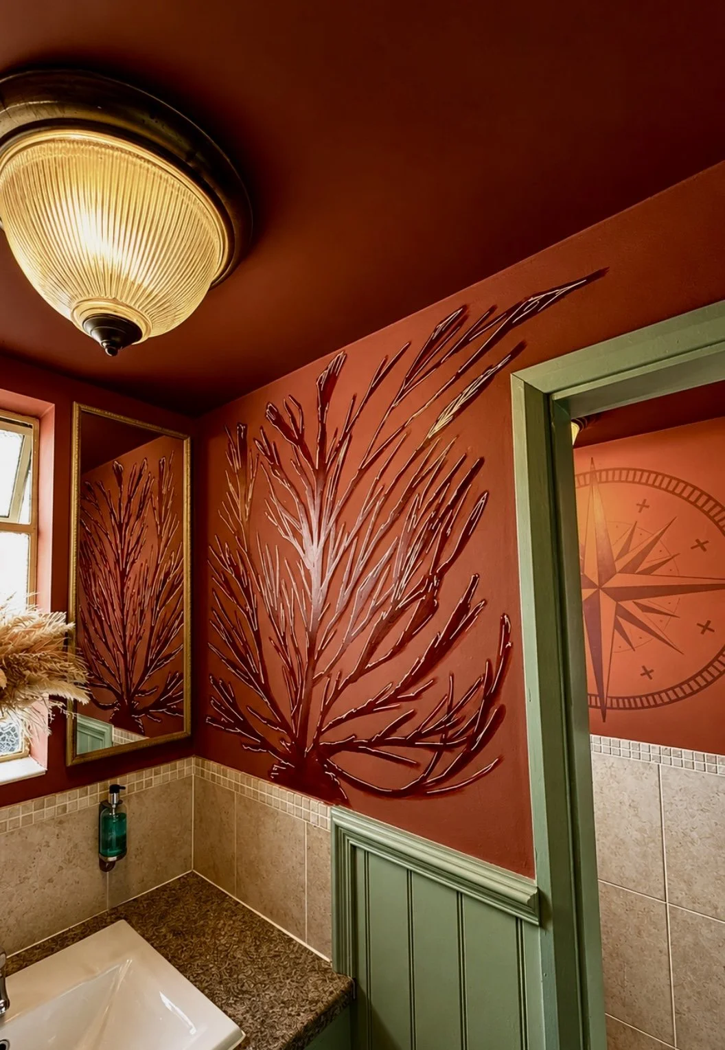 Interior bathroom with a ceiling light fixture, a mirror, a green hand soap dispenser, and a wall decorated with painted branch and leaf designs, and a ceiling with a compass rose pattern.