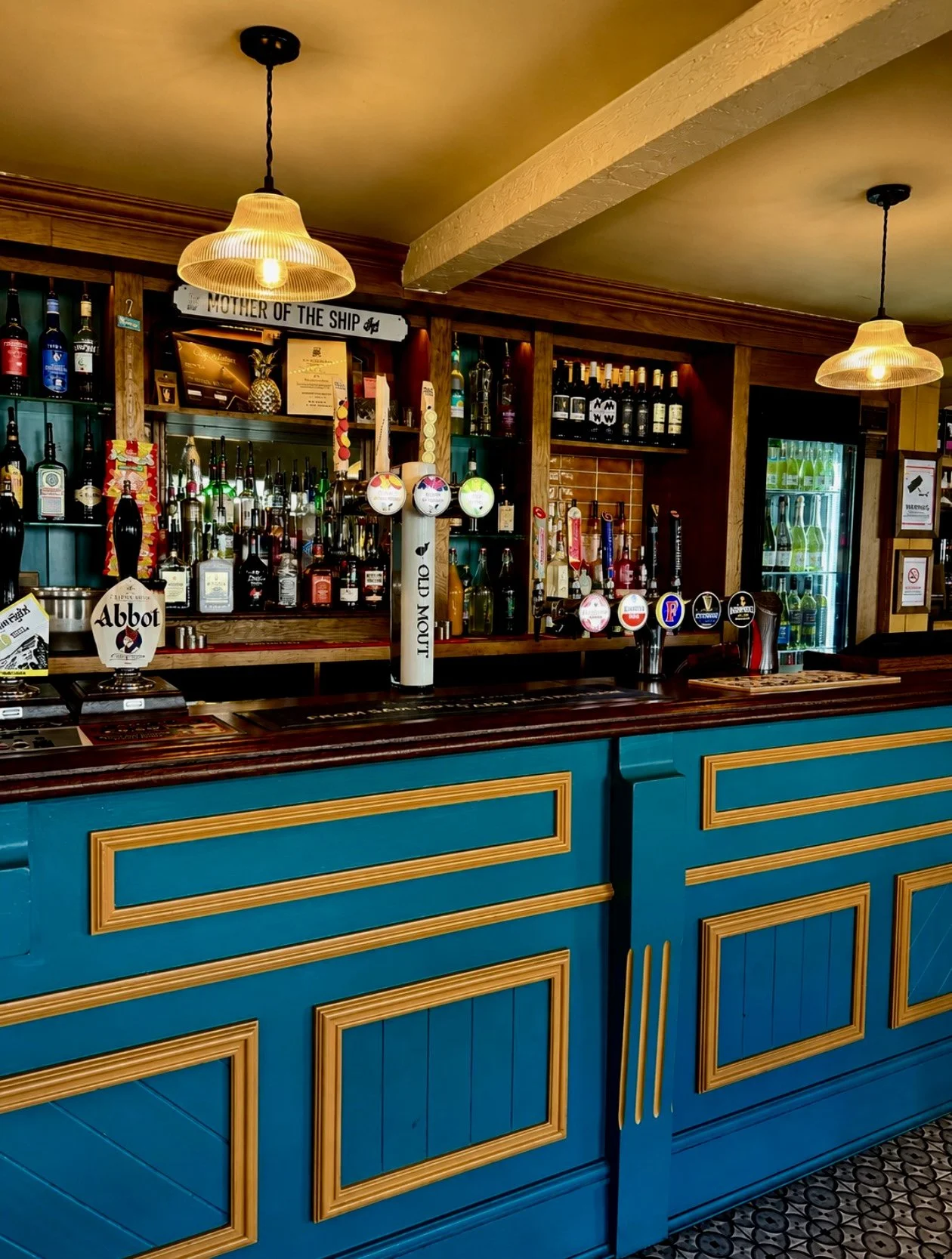 View of a bar with a blue and gold counter, stocked with various liquor bottles and beer taps, warm lighting, and a refrigerator with bottled drinks in the background.