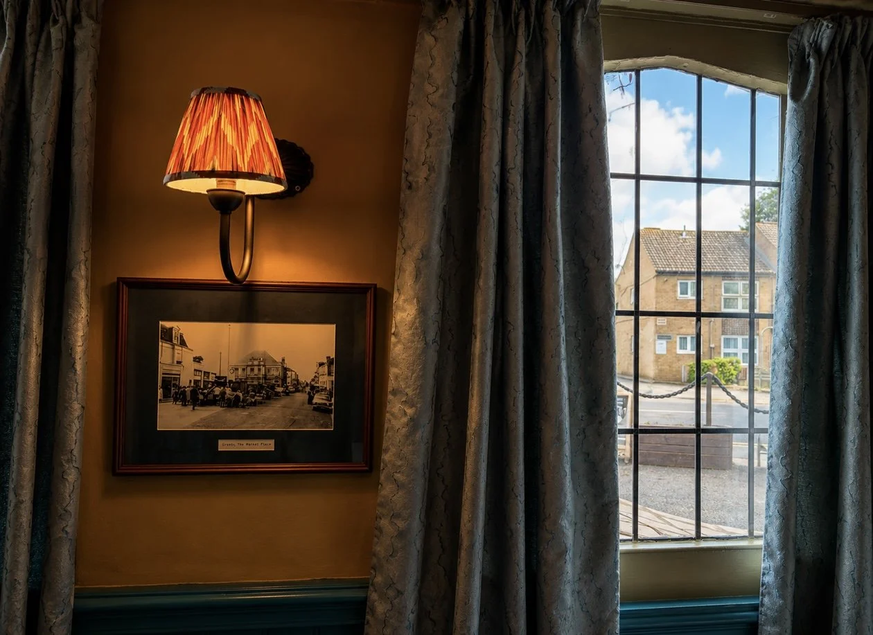 Interior view of a cozy room with a vintage photograph on the wall, a wall-mounted lamp with a red patterned shade, and a large window with patterned curtains showing a sunny street scene with houses outside.