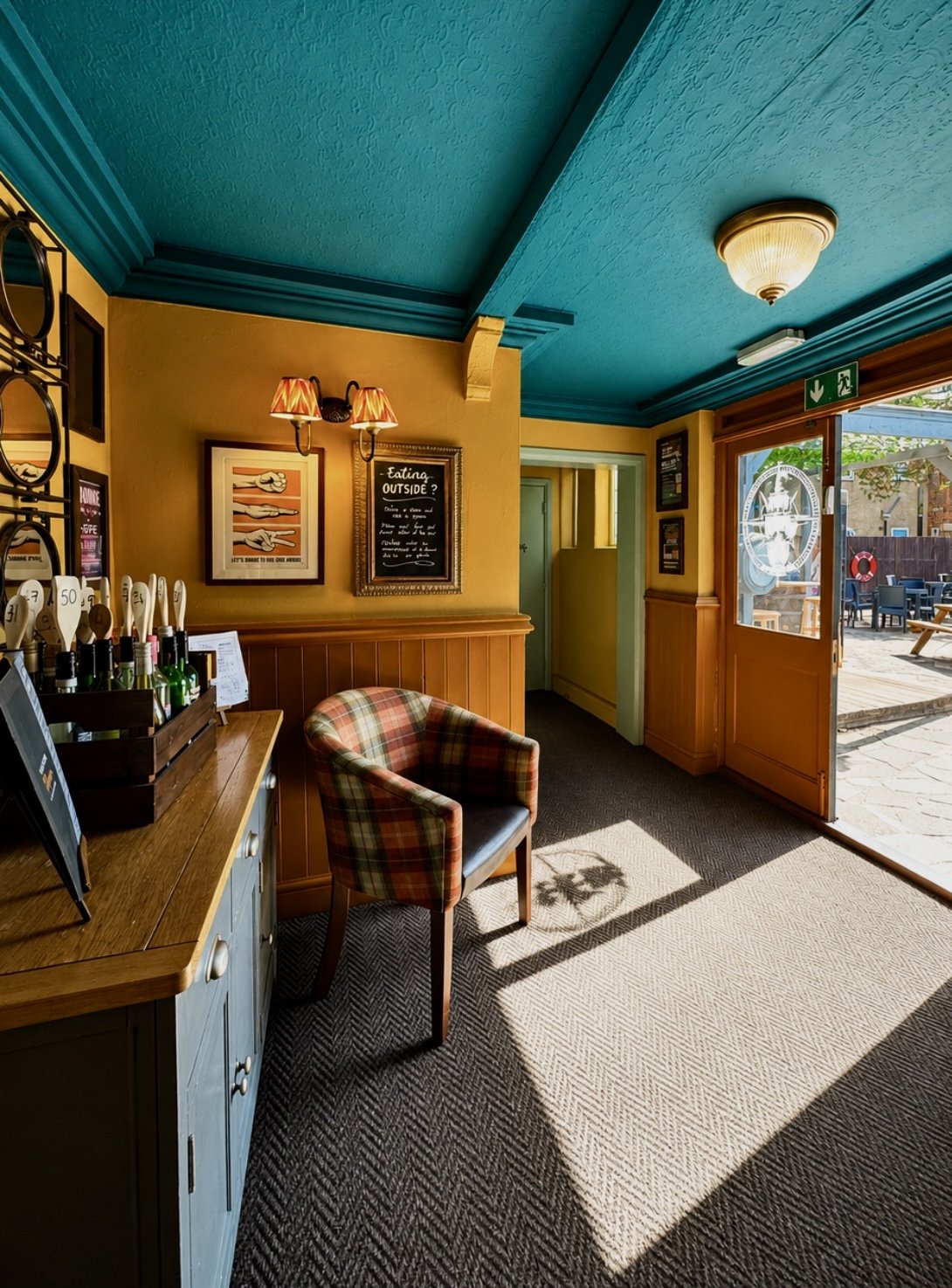 Interior of a cozy cafe with a plaid upholstered chair, a wooden counter with bottles and beer taps, framed pictures and a chalkboard on yellow walls, blue ceiling, and an open door leading outside with outdoor seating visible.