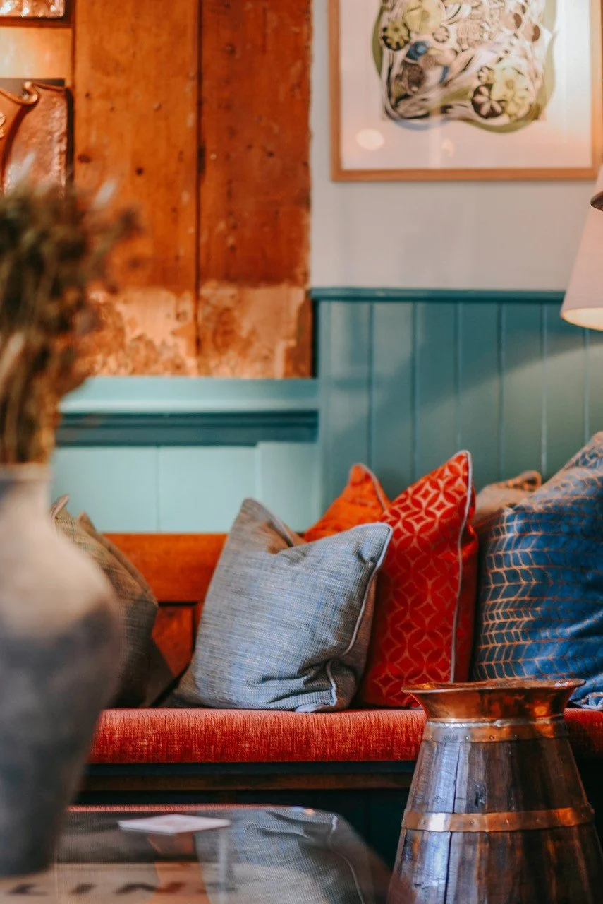 A cozy living room with a teal wooden-paneled wall, a reddish-orange couch with multiple decorative pillows, a copper vase on a table, and framed artwork on the wall.