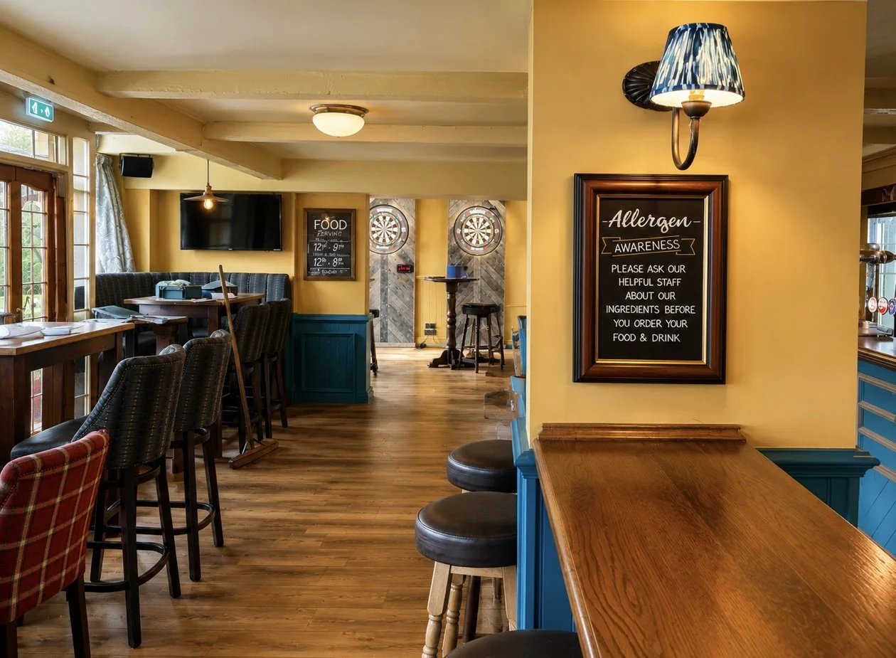 Interior of a cozy restaurant or pub with wooden floors, yellow walls, and a mix of bar stools and tables. There are dartboards in the back, a flat-screen TV, and a chalkboard menu. A framed allergen warning sign hangs on the wall.