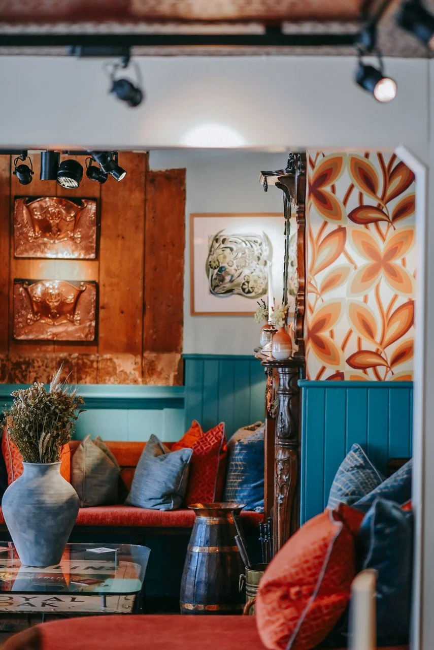 A cozy living room corner with colorful pillows on a red sofa, a large vase with dried flowers, patterned wallpaper, wooden accents, and wall art.