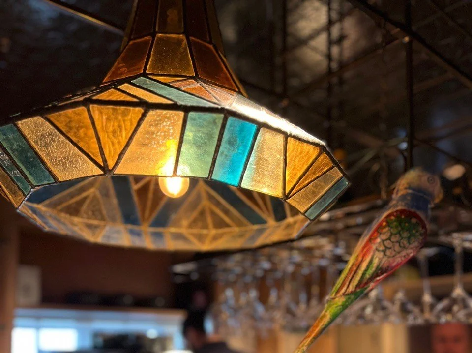 Close-up of a stained glass pendant lamp with a warm light, hanging in a dimly lit room. A colorful parrot figurine is also visible in the background.