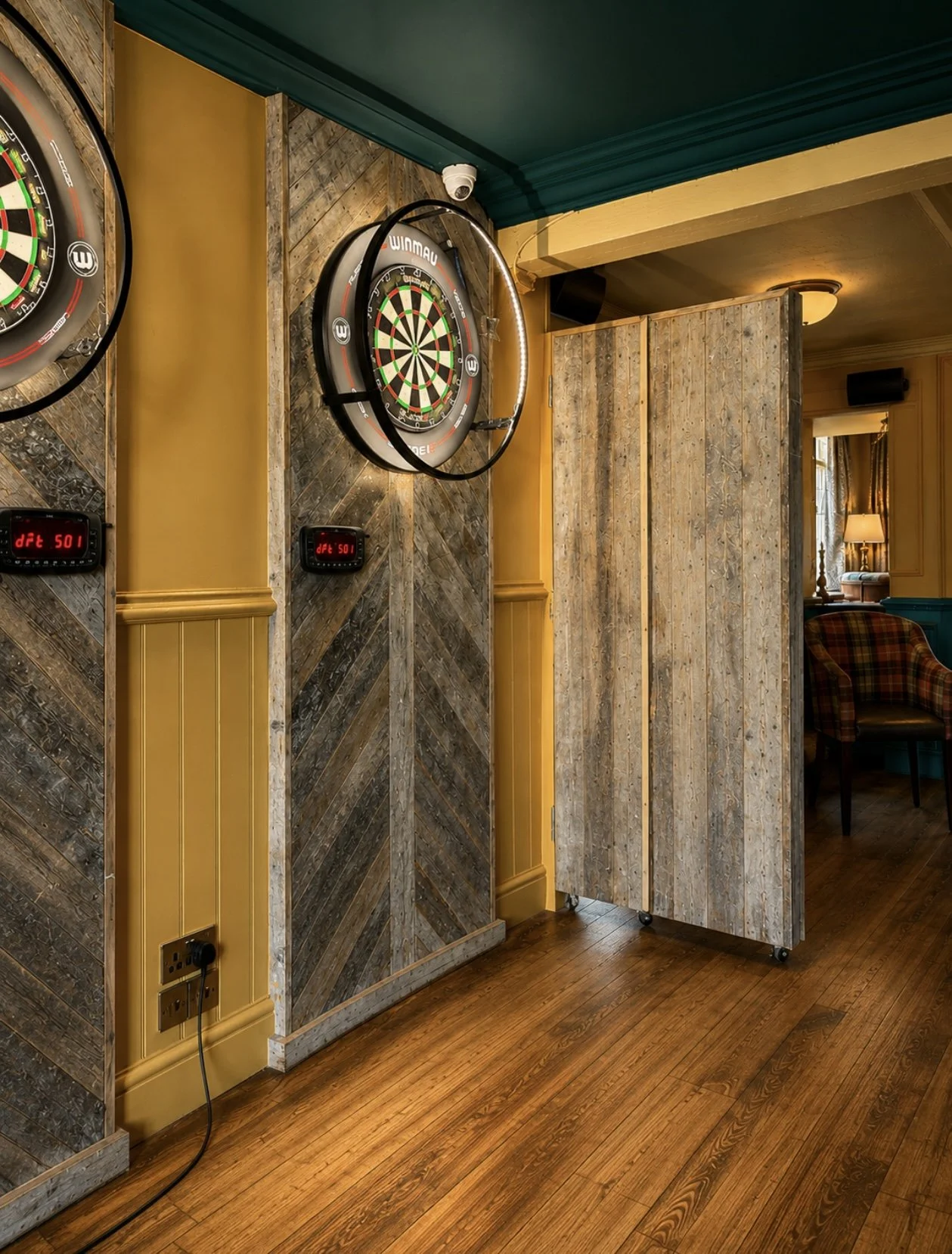 Two electronic dartboards mounted on a wooden wall with a rustic finish, in a cozy room with wooden floors and warm lighting. There is a wooden privacy screen partially covering the dartboards, and a lounge area with a chair and a lamp is visible in 
