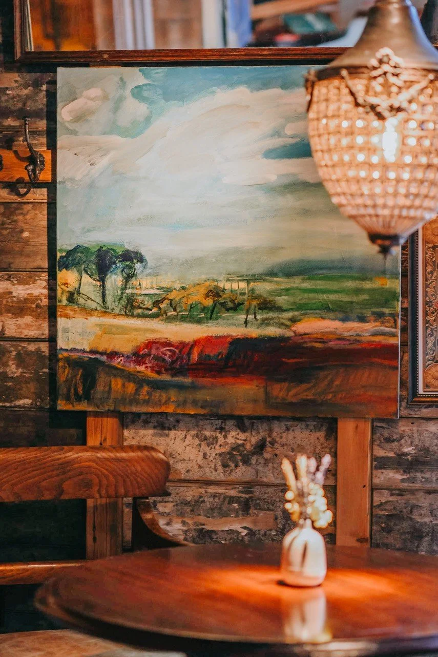 A landscape painting depicting fields, trees, and a cloudy sky, hung on a rustic wooden wall in a cozy interior with warm lighting.