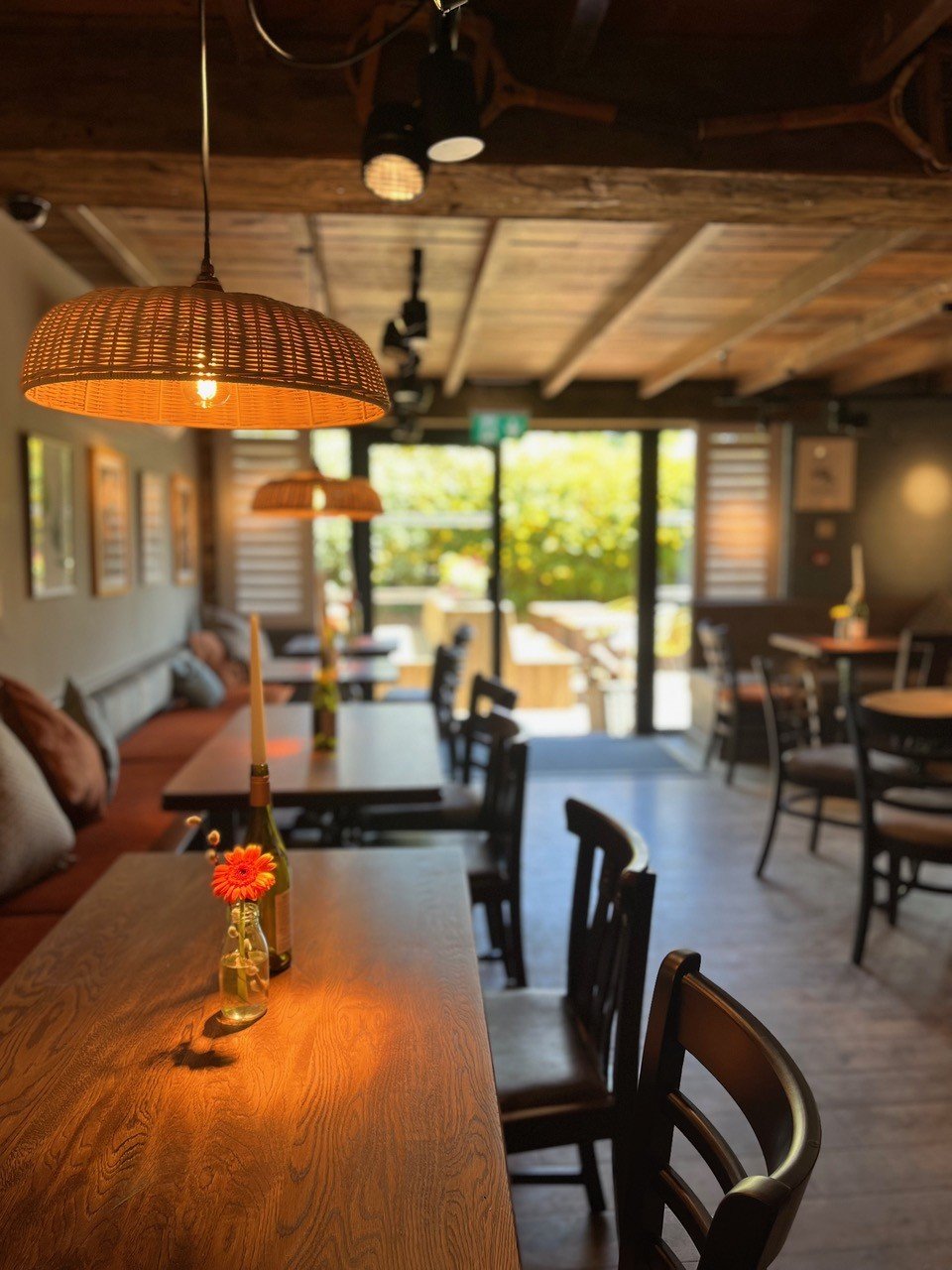 A cozy restaurant interior with wooden furniture, hanging wicker lamps, and small vases with flowers on the tables, with a view of the outdoor patio through large glass doors.