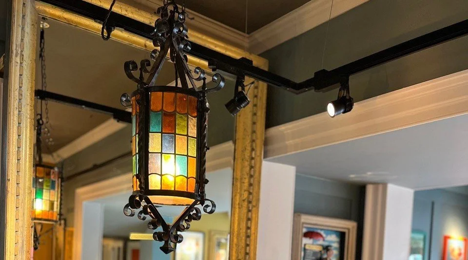 A hanging stained glass lantern lamp with a metal frame in a gallery or art space with framed artworks and a large mirror reflecting some of the room.