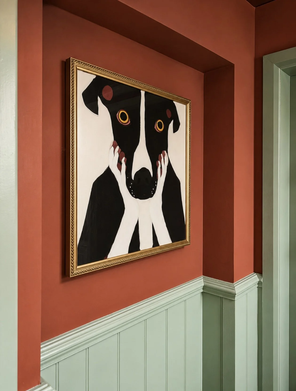 Painting of a black and white dog with ginger-colored eyes, holding its face with paws, framed and hanging on a red wall in a room with green wainscoting.