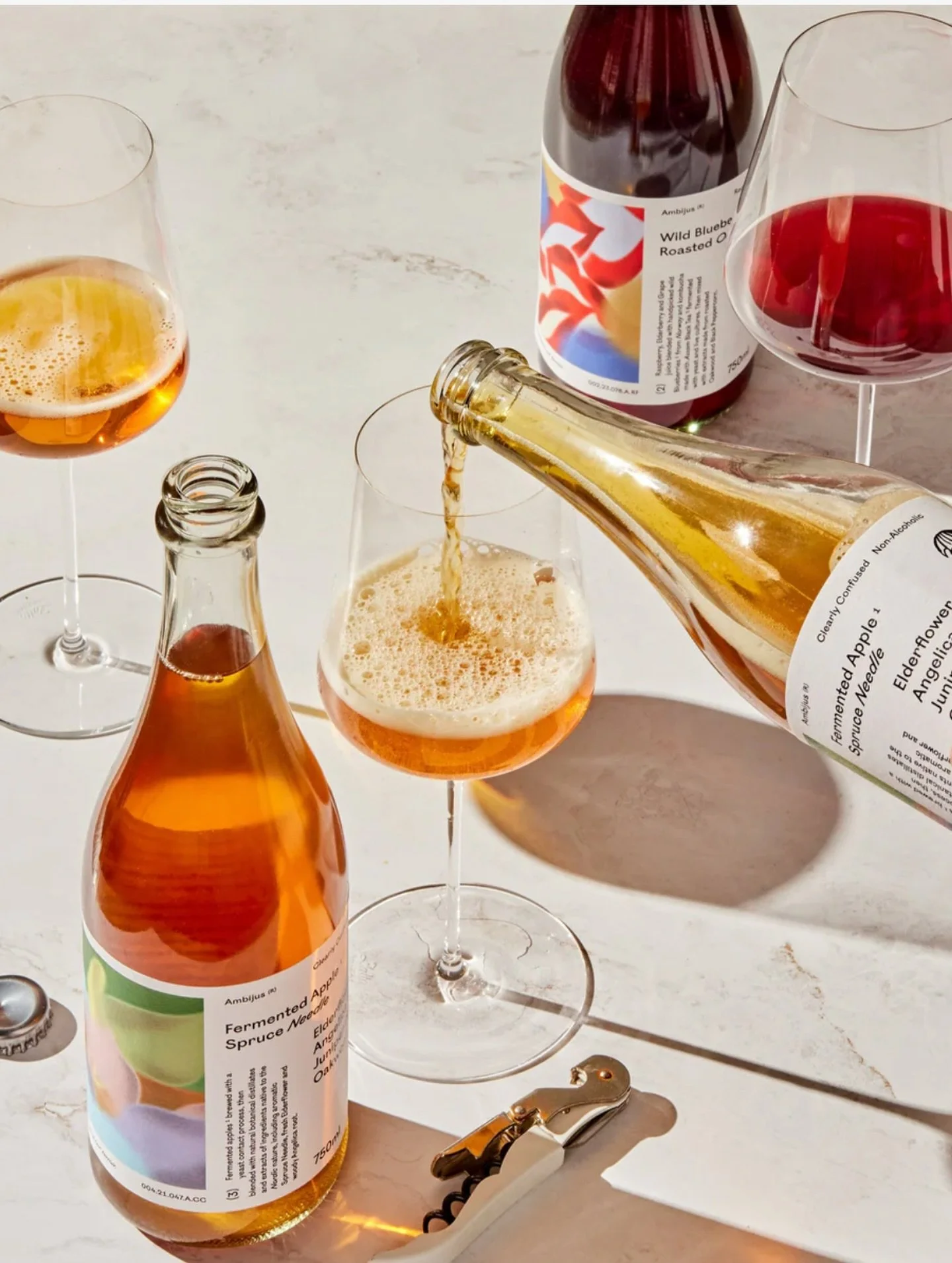 Et glass med en brunsvart drikke som blir fyllt fra en flaske med en merket etikett, omgitt av flere tomme glass og en flaske uten etikett, på et lyst bord med et verktøy og en hvit overflate.