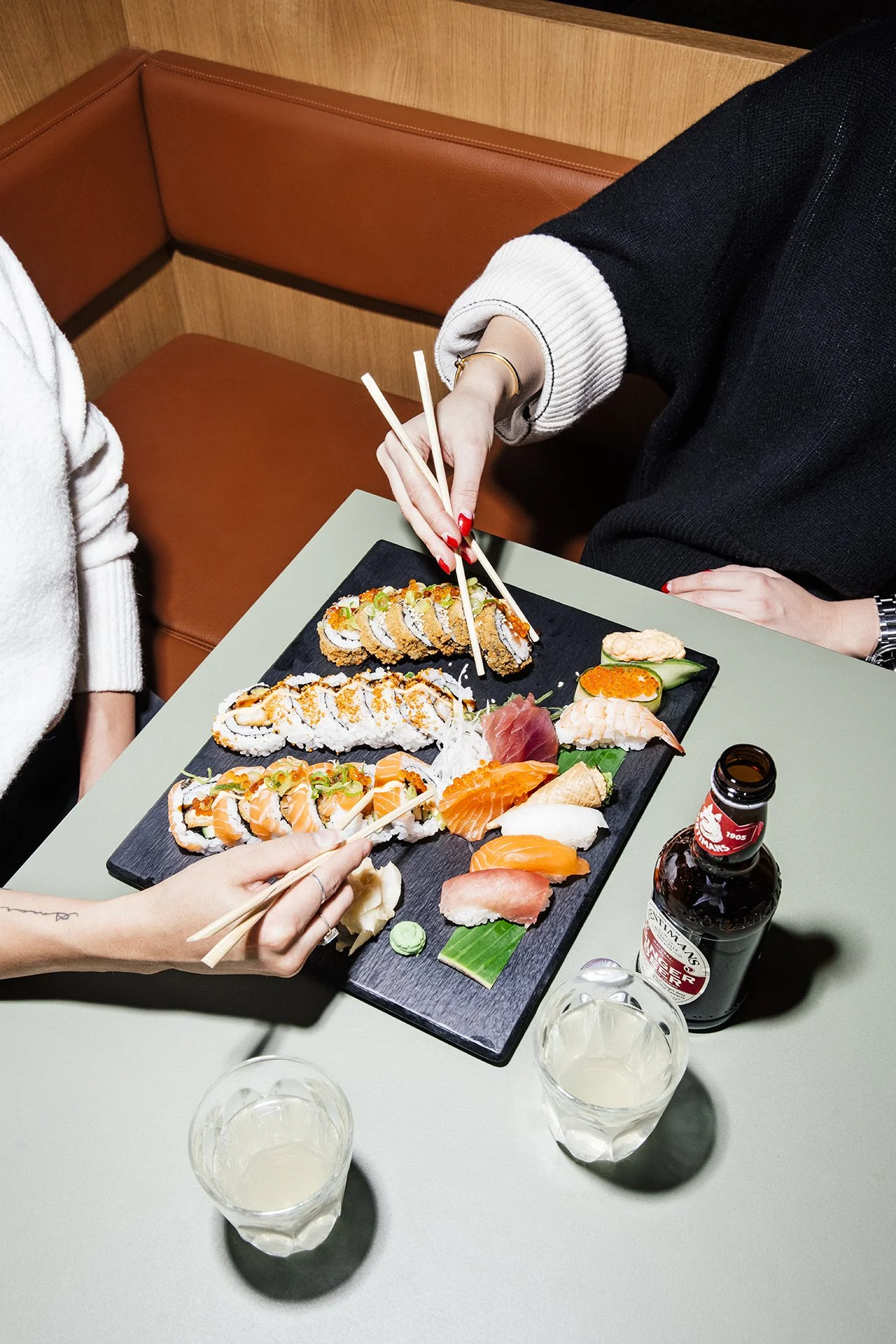 Sushi på en svart serveringsplate med mennesker som spiser med pinner, alkoholholdige drikker og en ølflaske på bordet.