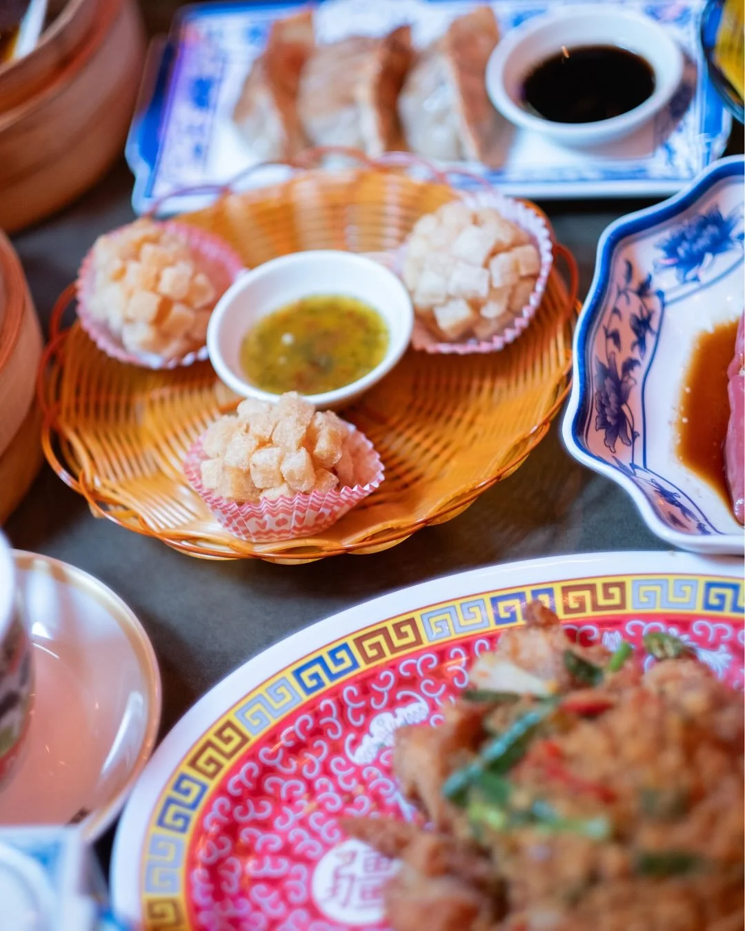 Bildet viser en flettet wok med krydret saus og grønnsaker, omgitt av tradisjonelle kinesiske retter og dim sum med saus, på fargerike tallerkener.