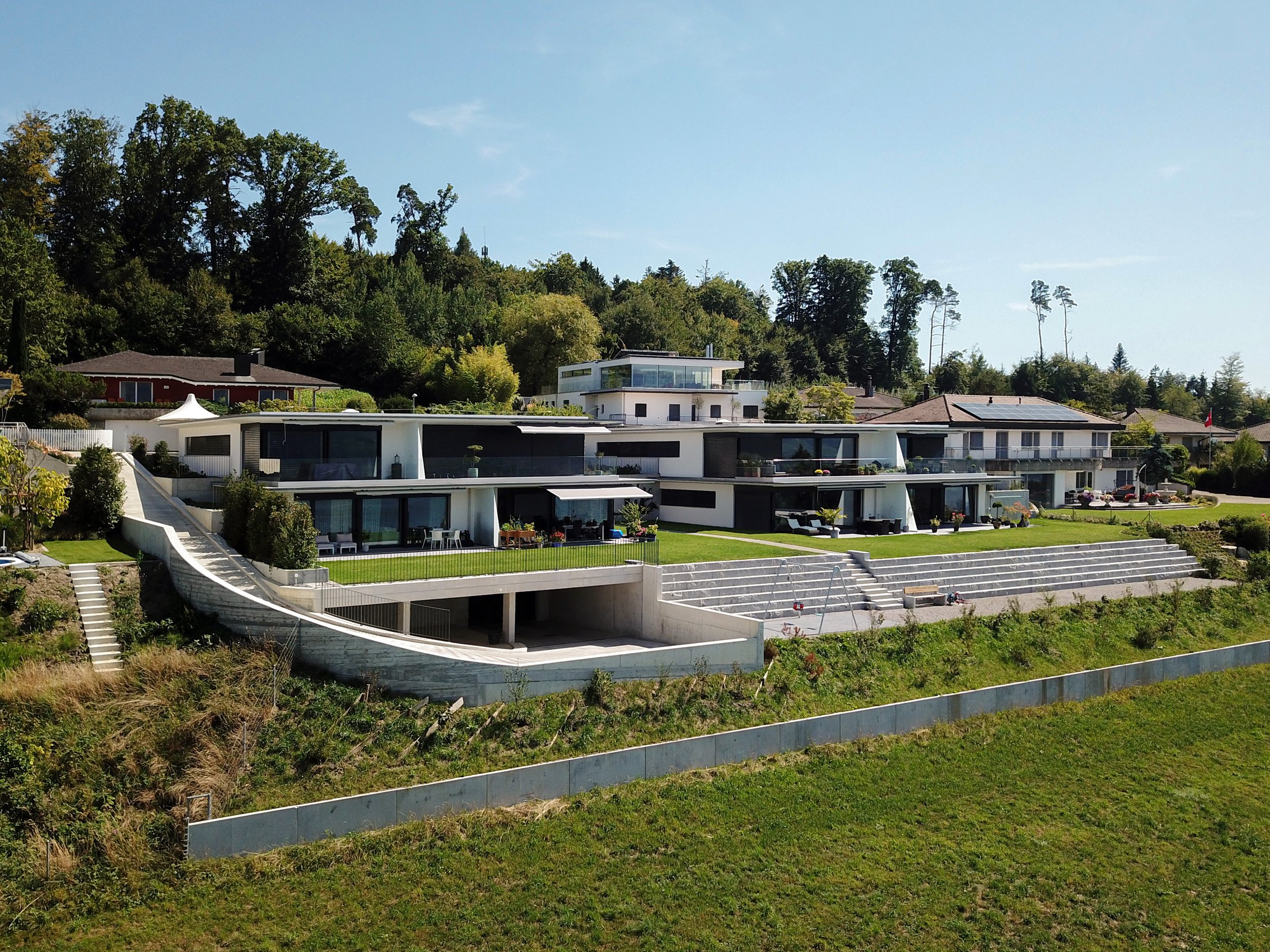 Moderne weiße Häuser mit großen Fensterflächen und Gartenterrassen, vor einem bewaldeten Hügel und einem klaren blauen Himmel.