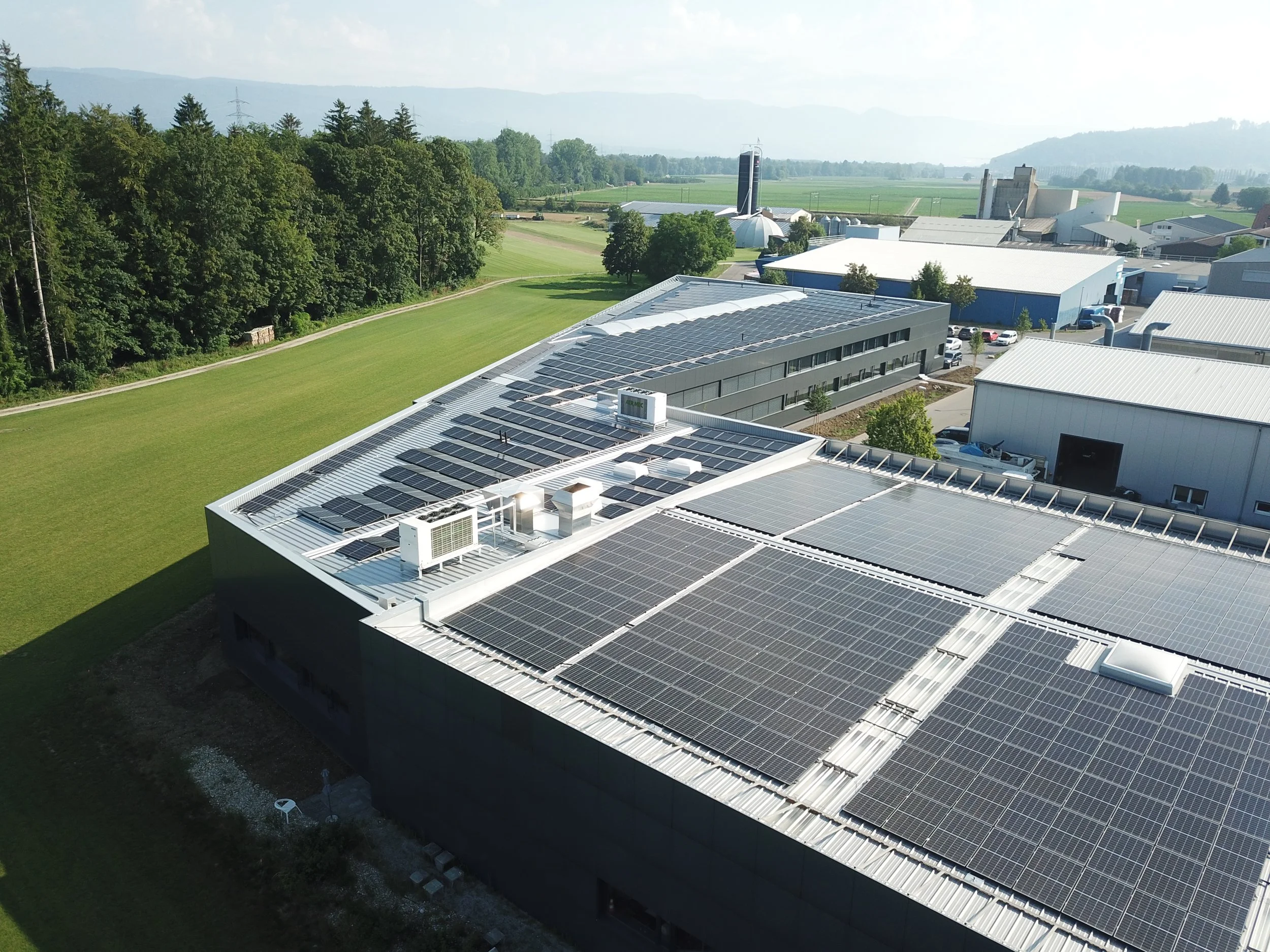 Ein Gebäude mit Solarpanelen auf dem Dach, umgeben von grünem Rasen und Bäumen, mit landwirtschaftlichen Flächen im Hintergrund.