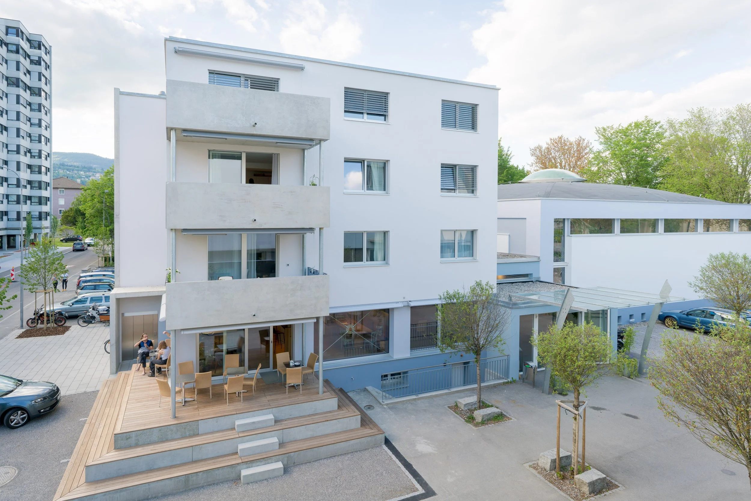 Moderne weiße mehrstöckige Wohngebäude mit Balkonen und großen Fenstern, im Vordergrund ein Terrassendeck mit Sitzgelegenheiten und Tischen, daneben ein parkender PKW, umgeben von Bäumen und einem Parkplatz im Hintergrund.