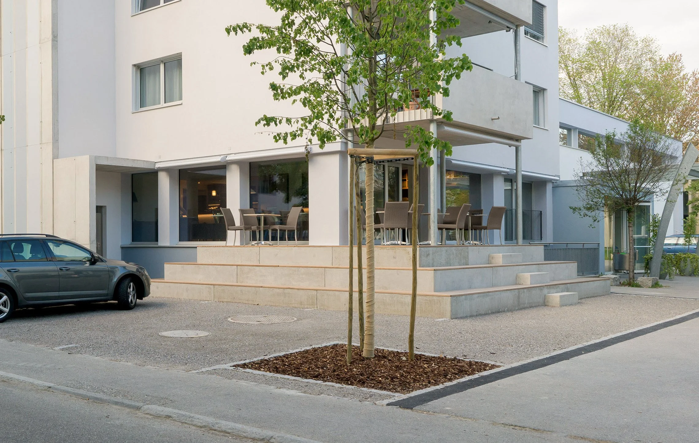 Mehrstöckiges weißes Gebäude mit großen Fenstern und Balkon, im Vordergrund sind ein Baum und eine Terrasse mit Stühlen und Tischen, im Hintergrund sind weitere Bäume und ein grauer Himmel.