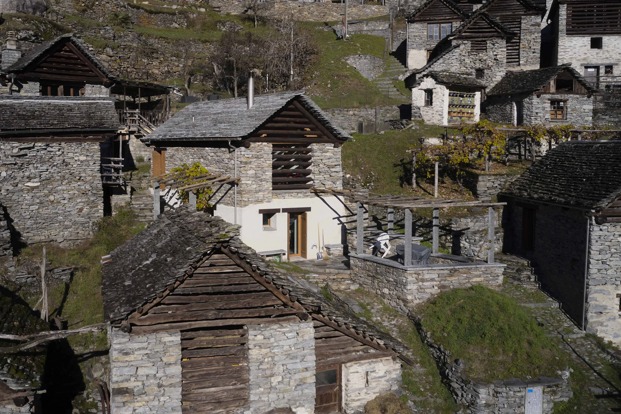 Dieses Bild zeigt eine Ansammlung von traditionellen Steinhäusern mit Holzdächern, eingebettet in eine hügelige Landschaft. Einige Häuser haben Balkone und kleine Gärten, während im Hintergrund terrassierte Stufen die Anhöhe erklimmen.