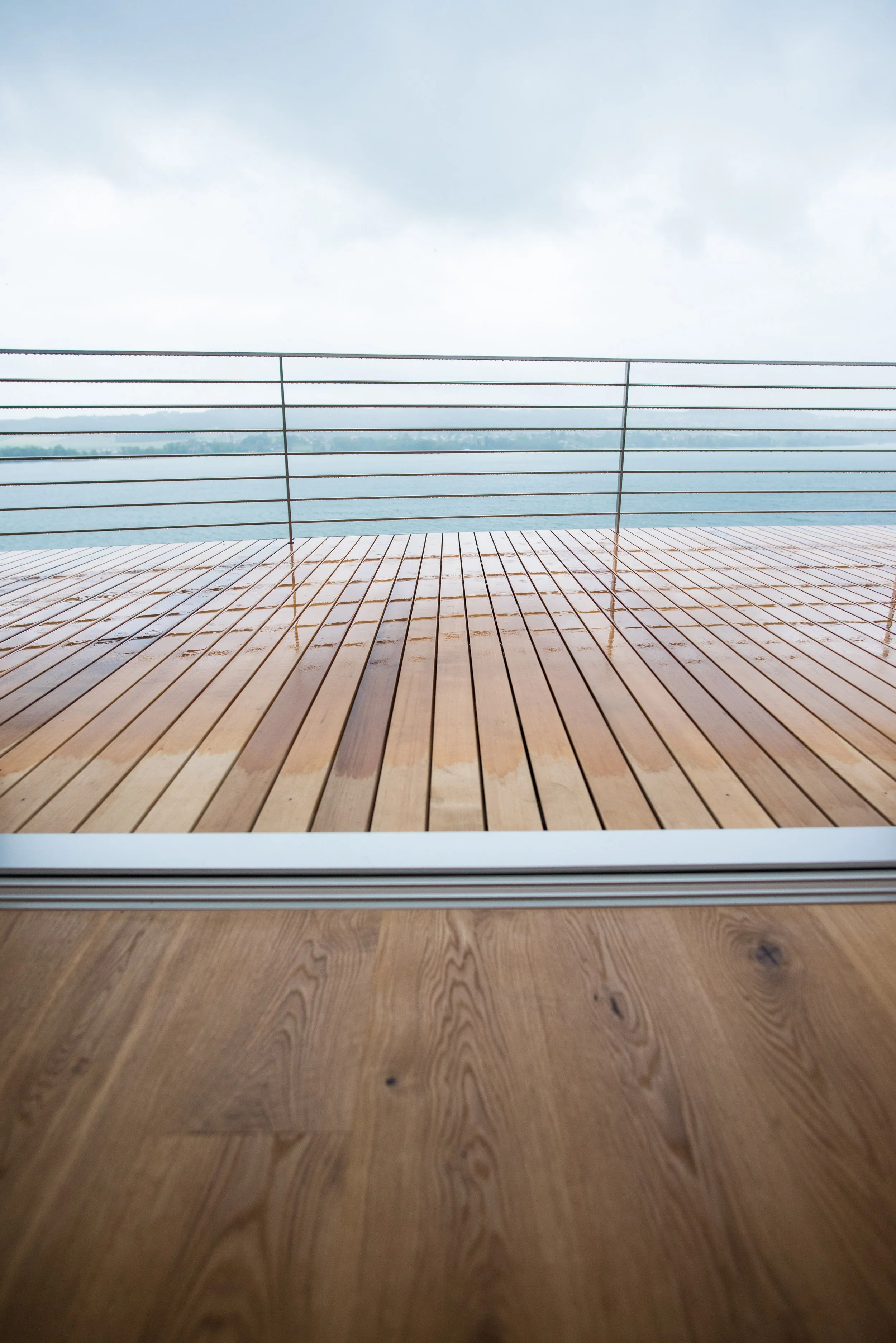 Blick durch eine Glasschiebetür auf eine Holzterrasse mit Wasser und Himmel im Hintergrund.