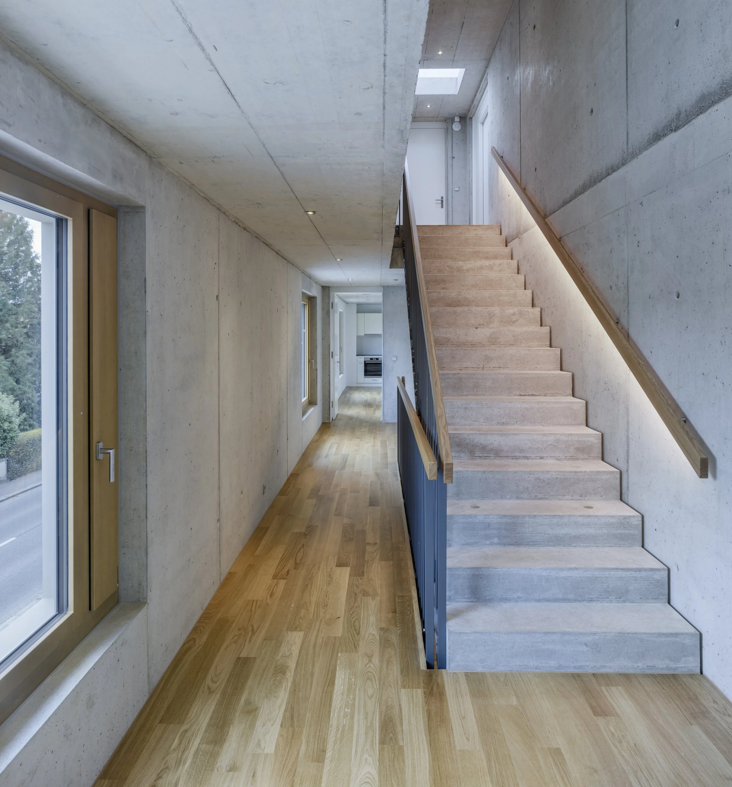 Innenansicht eines modernen Hauses mit Stufen aus Beton und Holz, große Fenster und helle Wände.
