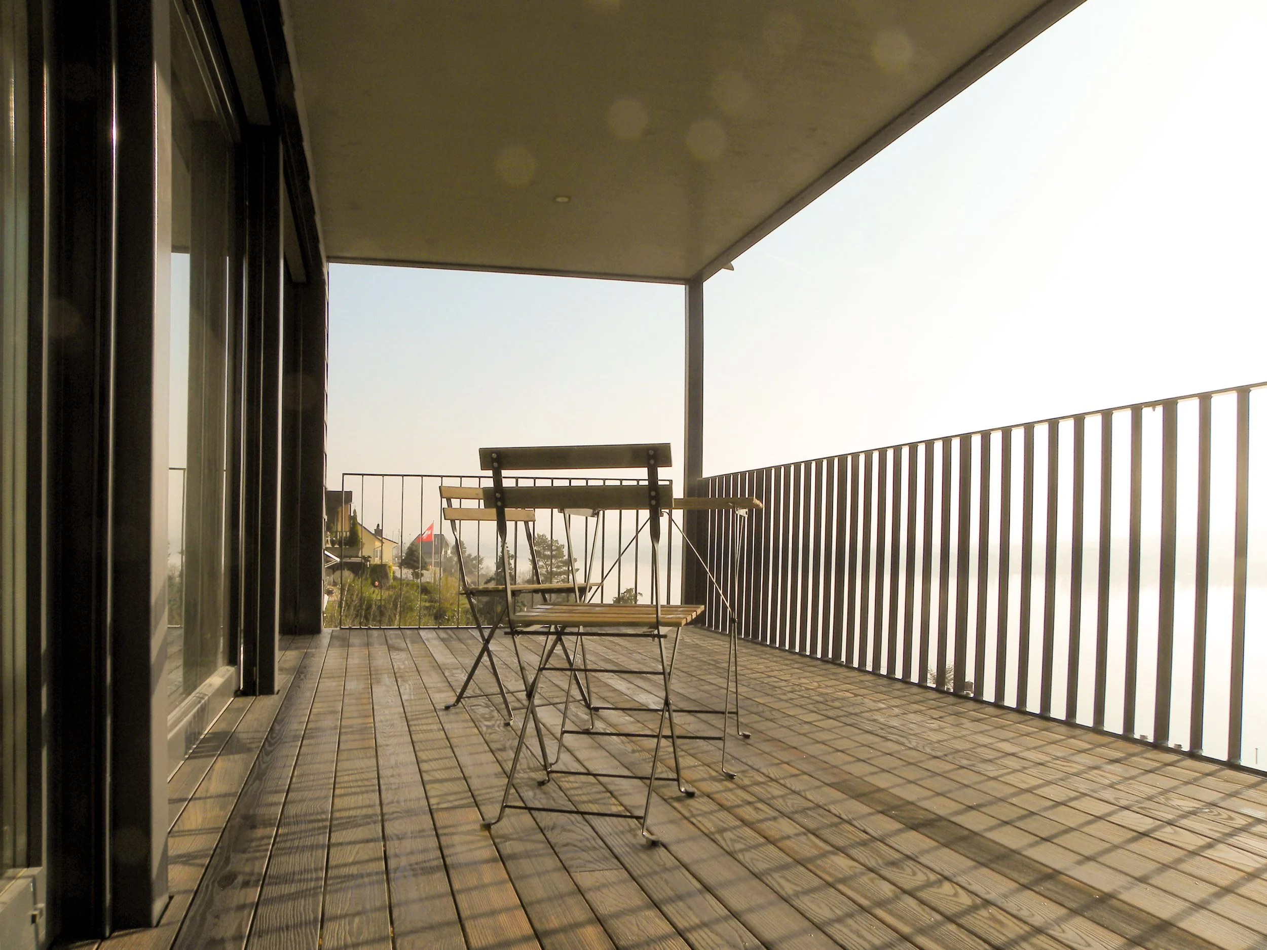 Ein Balkon mit Holzfußboden, einer weißen Metallbrüstung, einem kleinen Tisch und zwei Stühlen, Blick auf einen Himmel und wenige Gebäude im Hintergrund.