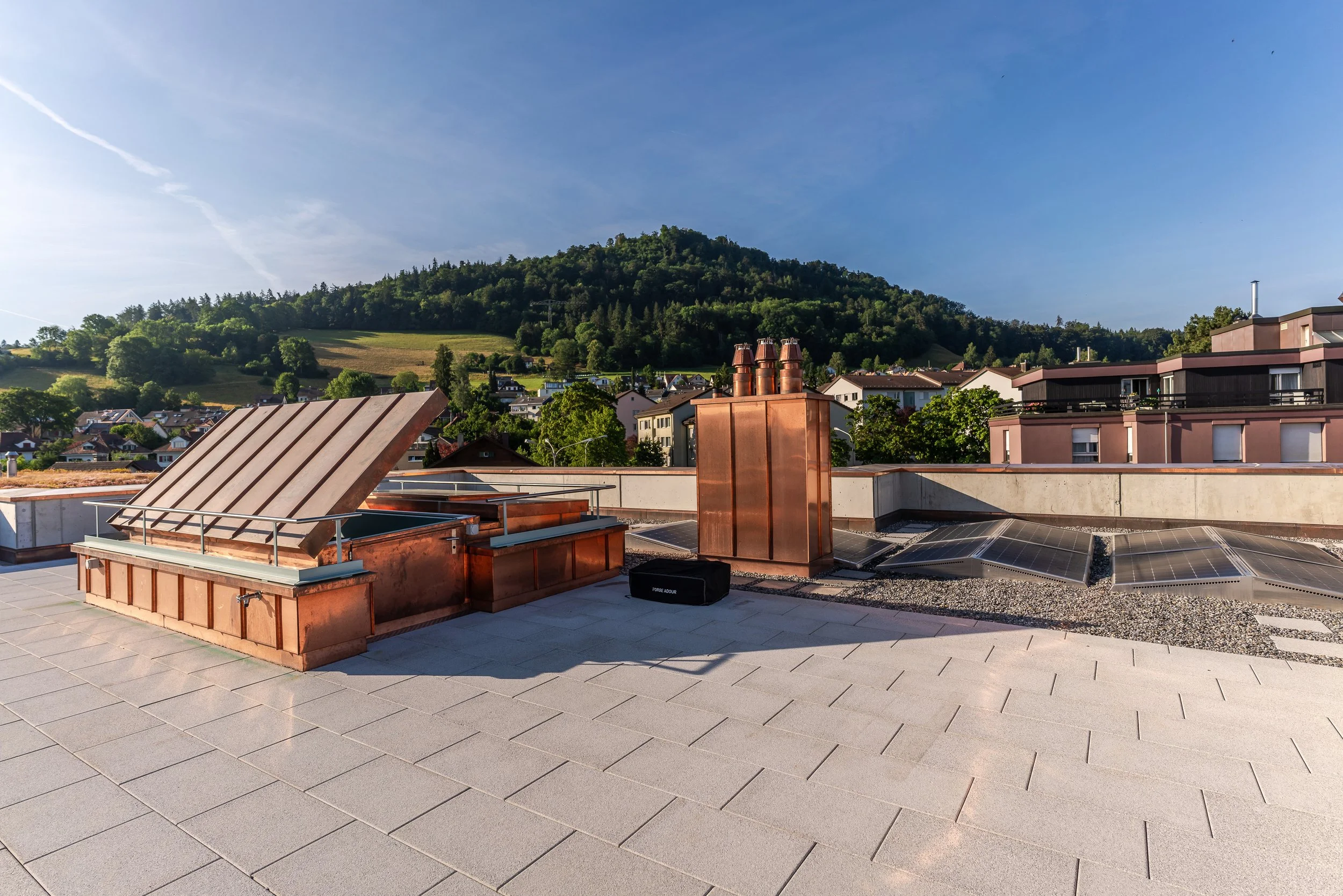 Dachterrasse mit Solarpaneelen, Kupferabdeckungen und Blick auf ein Hügellandschaft mit Häusern und Wäldern.