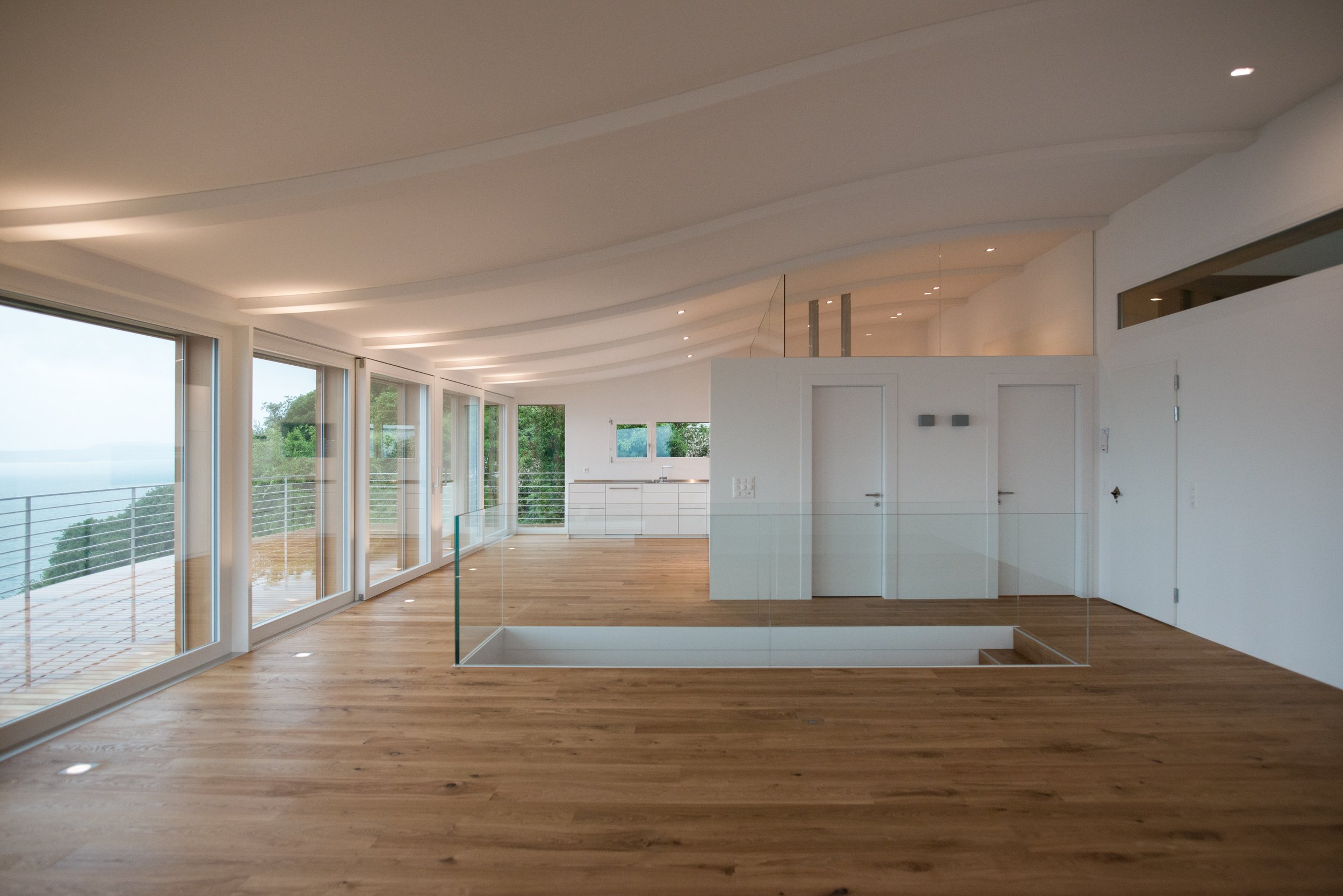 Ein moderner, heller Innenraum mit großen Fenstern, Holzboden und weißer Decke, der Blick auf eine grüne Natur im Hintergrund bietet.