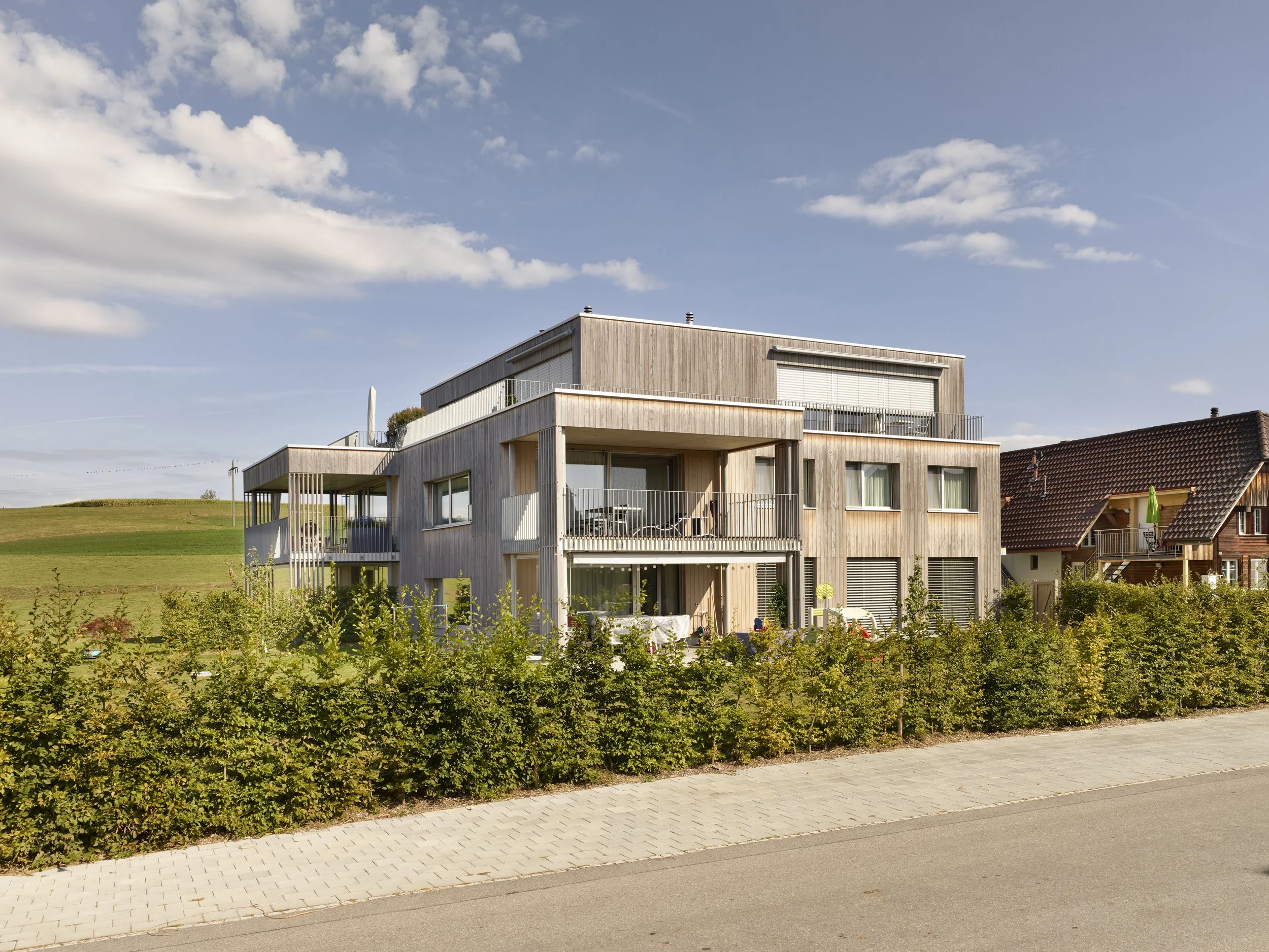 Modernes Gebäude mit mehreren Ebenen und Balkonen, umgeben von Büschen, bei sonnigem Himmel.
