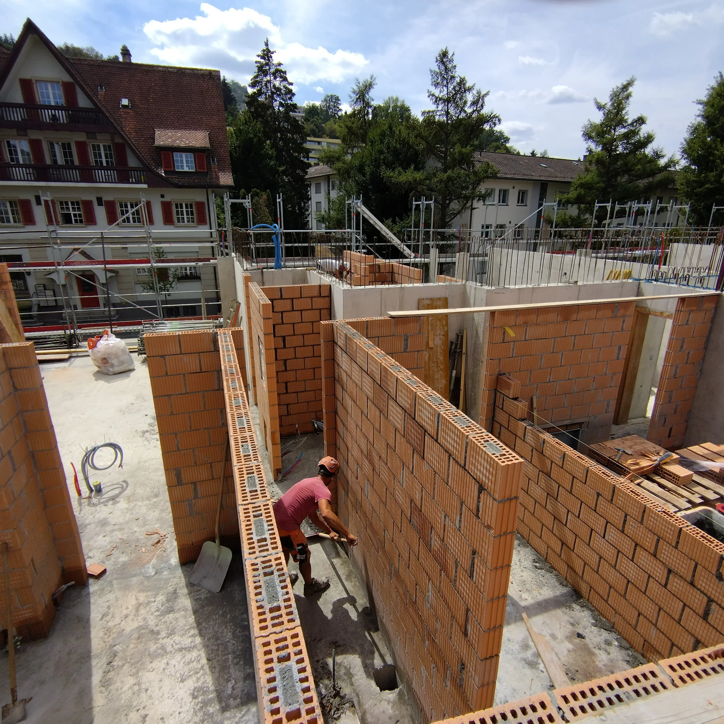 Bauarbeiter betreuen den Bau einer Hauswand aus Ziegelsteinen auf einer Baustelle vor einem Wohnhaus mit roten Dachziegeln und Bäumen im Hintergrund.