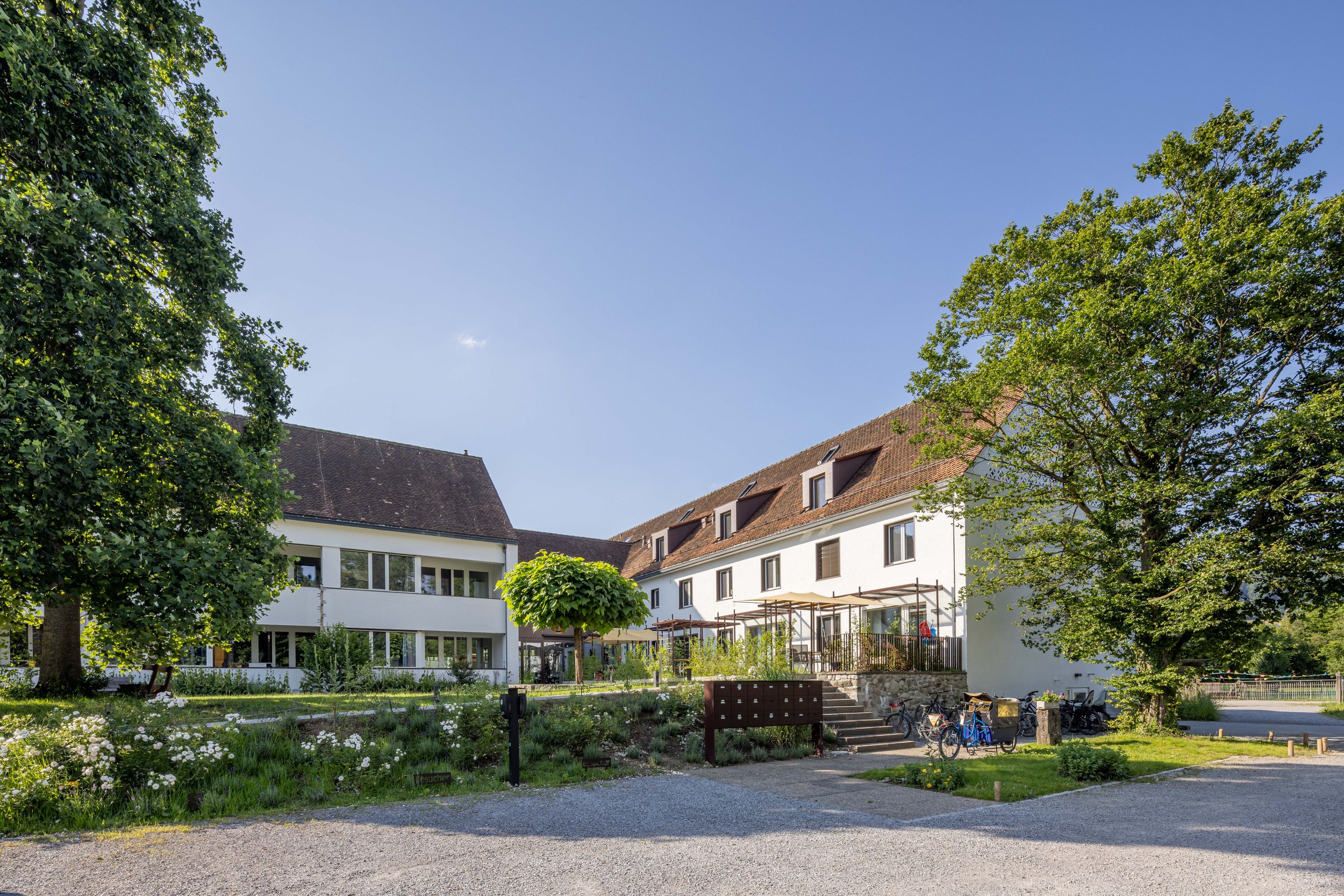 Grüner Garten mit Blumen, Bäumen, modernen weißen Gebäuden und Fahrrädern, bei sonnigem Himmel.