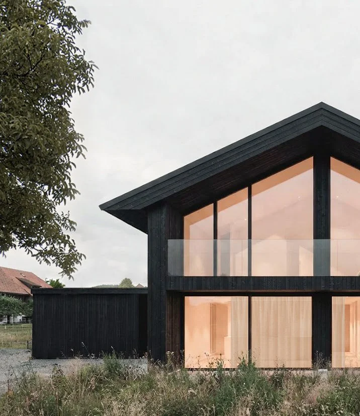 Modern zweistöckiges Haus mit schwarzen Holzfassaden, große Fenster und Balkon im Obergeschoss, umgeben von Natur mit Bäumen und Wiesen