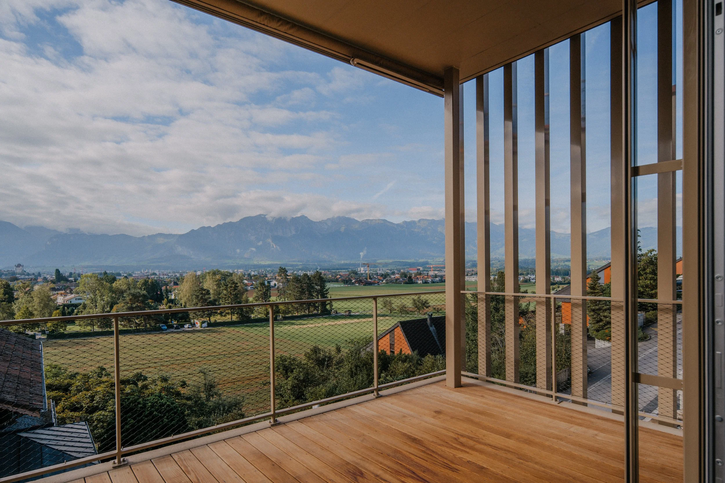 Balkon mit Holzdecke und Geländer, Blick auf eine grüne Landschaft mit Bäumen, Häuser und Berge im Hintergrund, bei sonnigem Himmel.