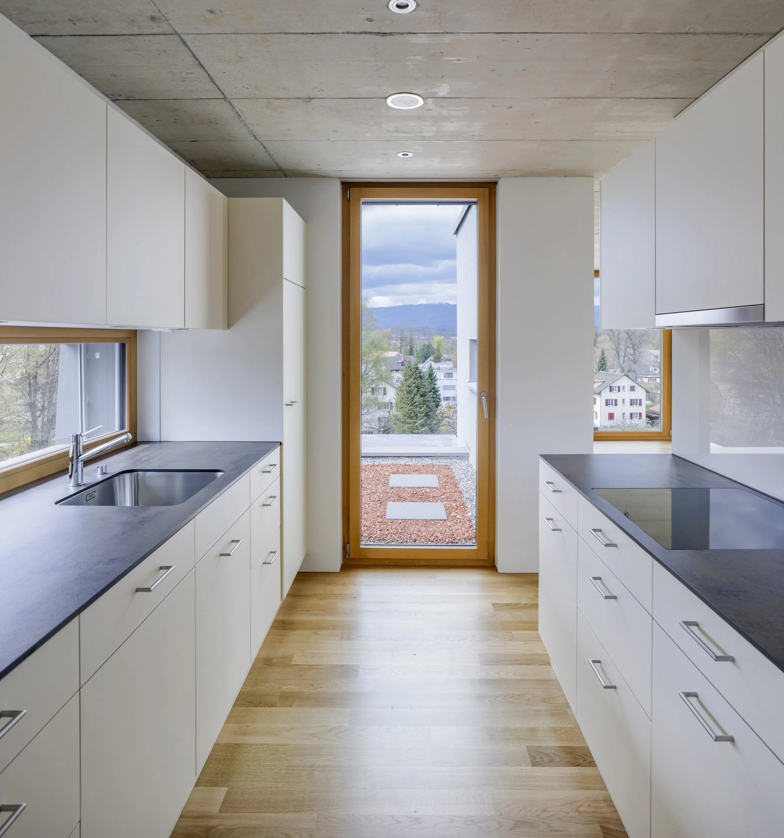 Moderne Küche mit weißen Schränken, grauen Arbeitsflächen und großen Fenstern mit Blick auf eine ländliche Landschaft.