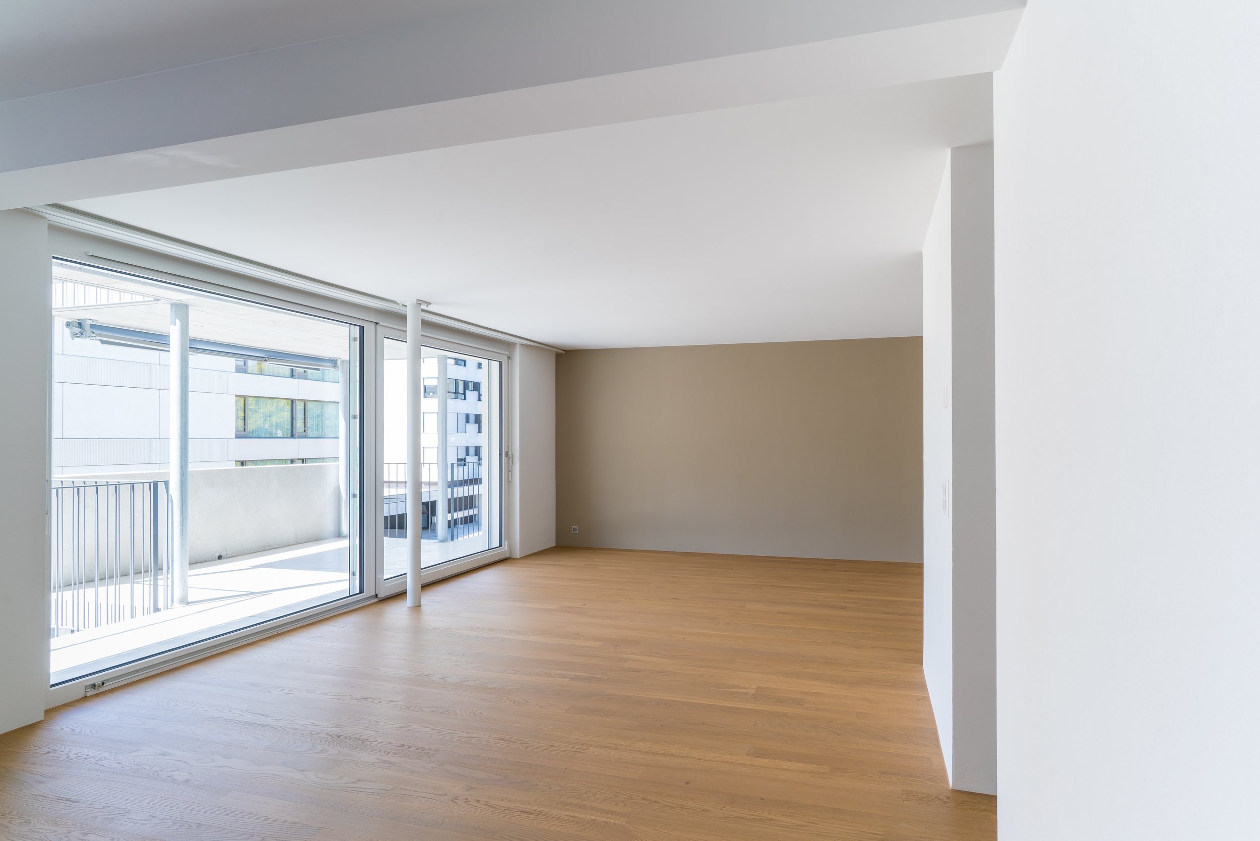 Leeres modernes Wohnzimmer mit großen Glastüren, durch die Sonnenlicht einfällt. Helle Wände und Holzboden schaffen eine freundliche Atmosphäre.