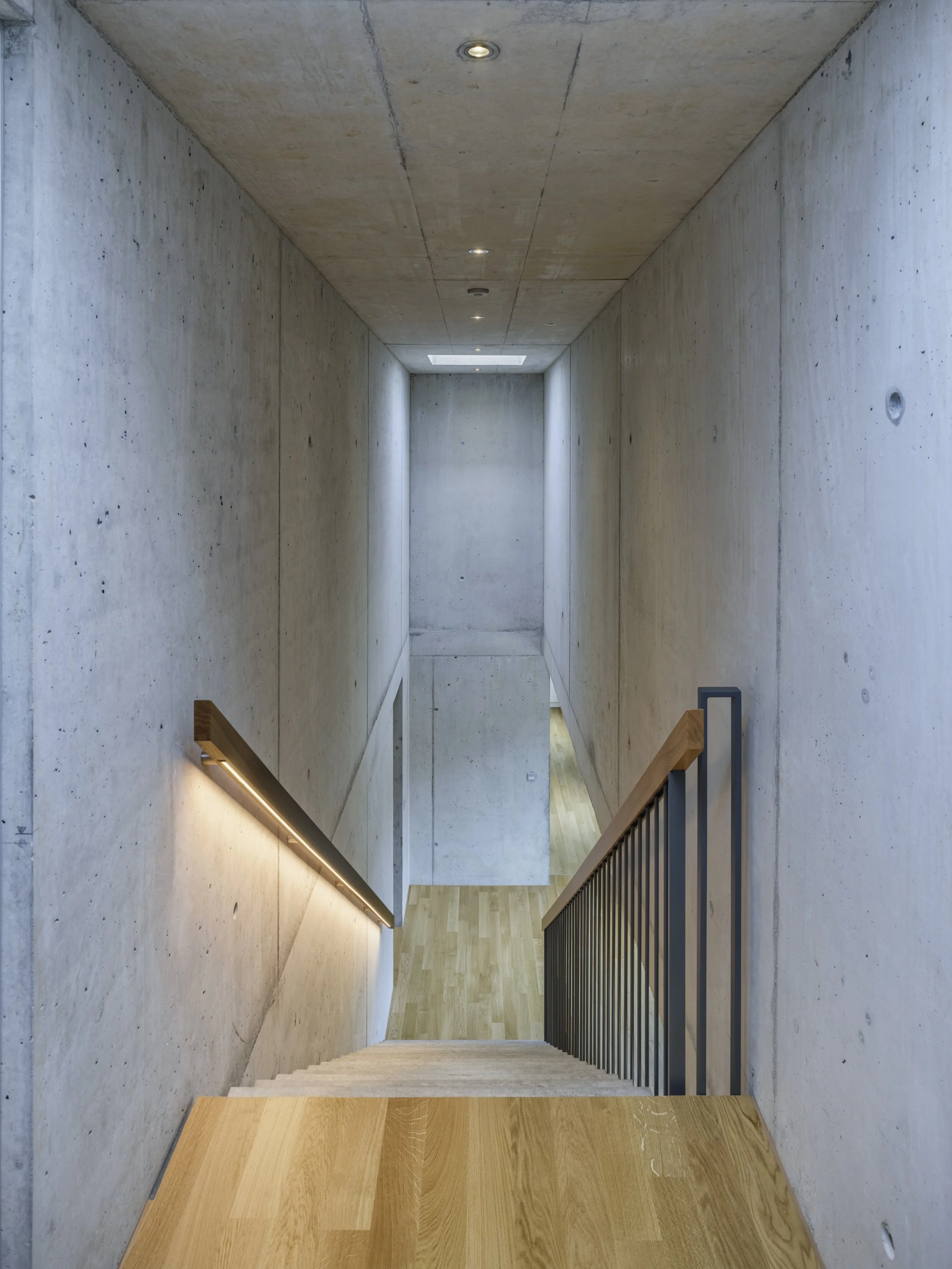 Innenaufnahme eines modernen Treppenhauses mit Betonwänden, Holzboden und einem schwarzen Metallhandlauf.