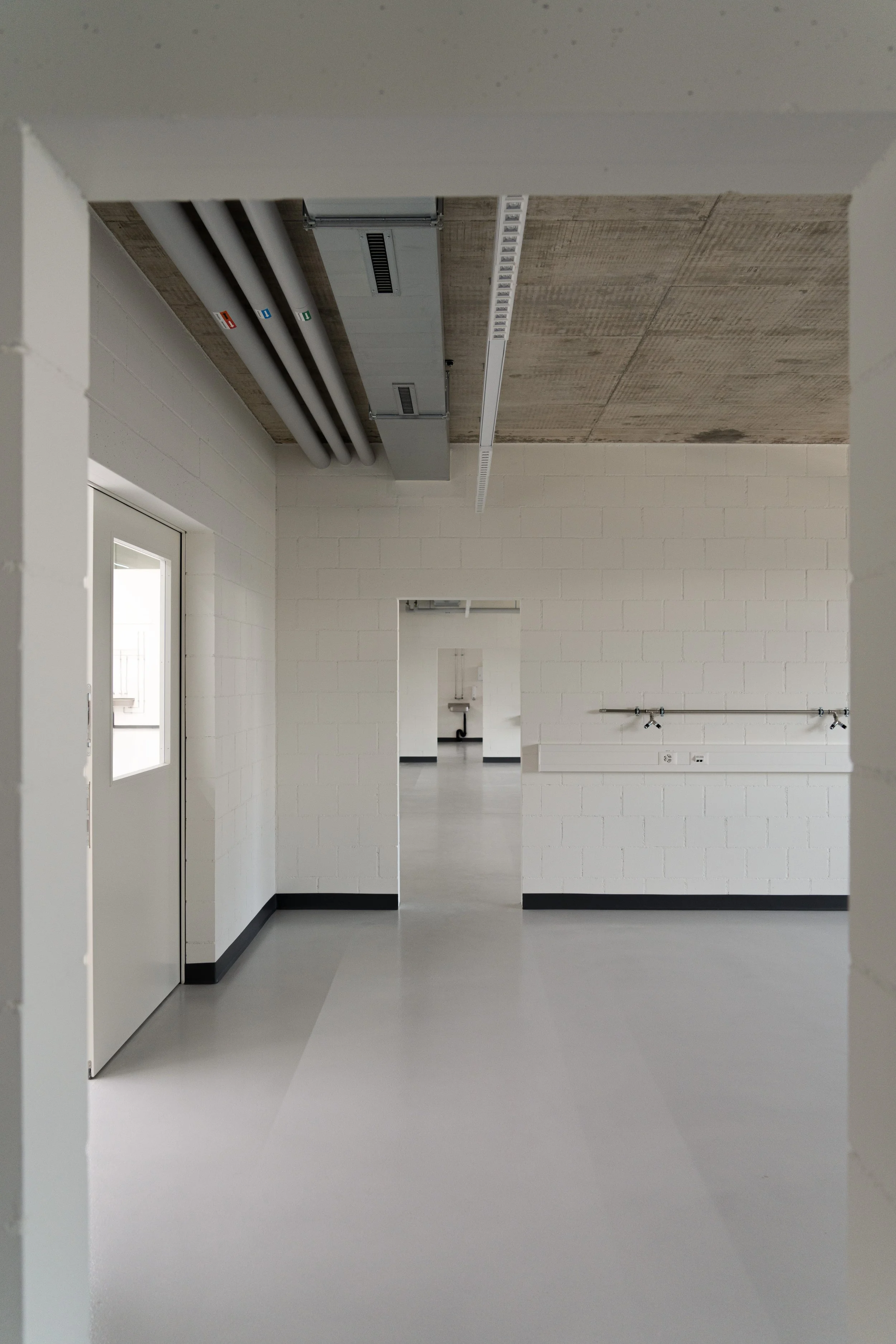 Leerer Flur in einem modernen Gebäude mit weißen Wänden, grauem Boden und einer Tür auf der linken Seite. An der Wand auf der rechten Seite sind Wasserhähne und Anschlüsse sichtbar.