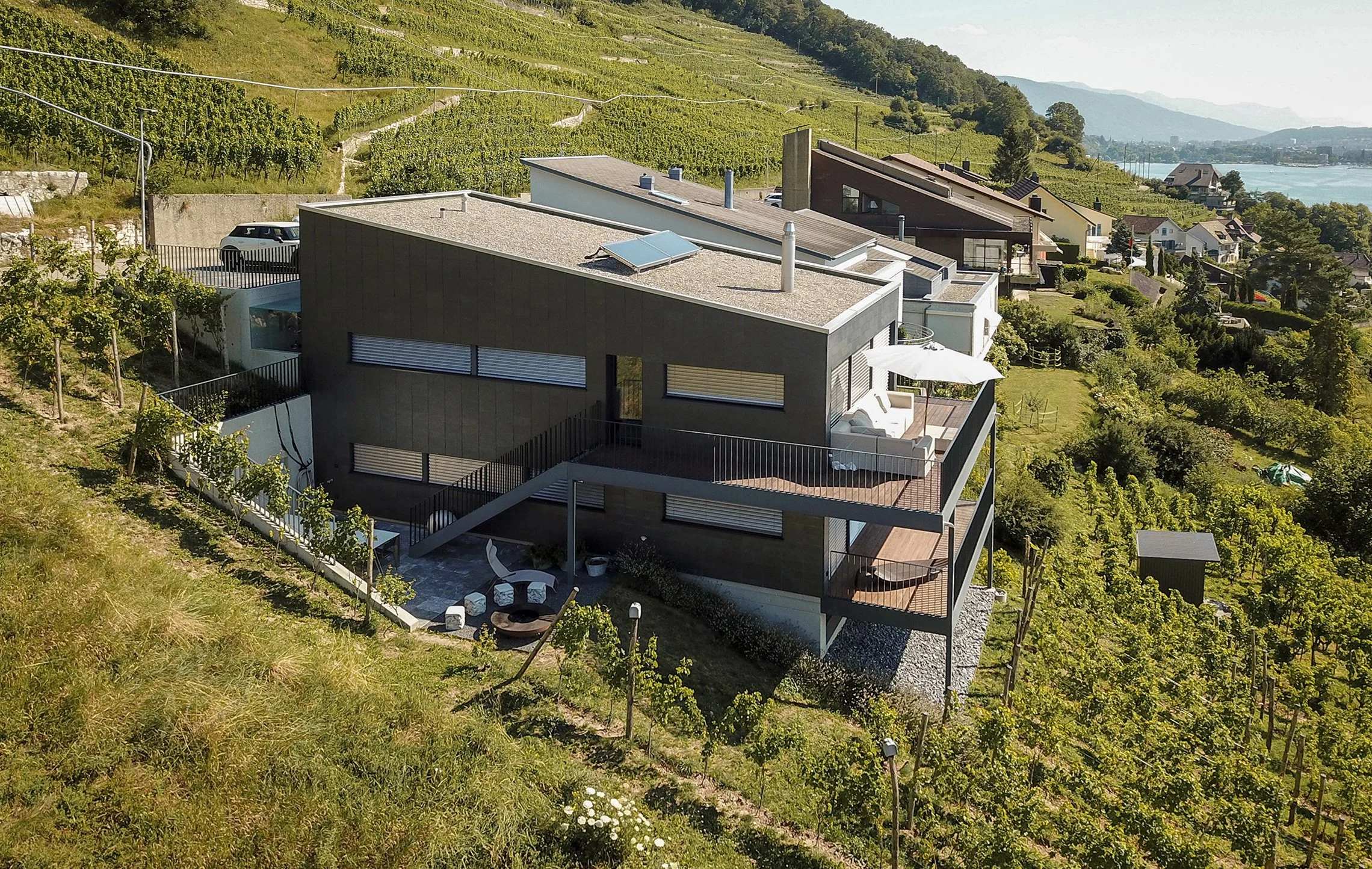 Moderne schwarze Villa auf einer hügeligen, terrassierten Landschaft mit Weingärten, in der Nähe eines Sees, umgeben von Bäumen und anderen Häusern.