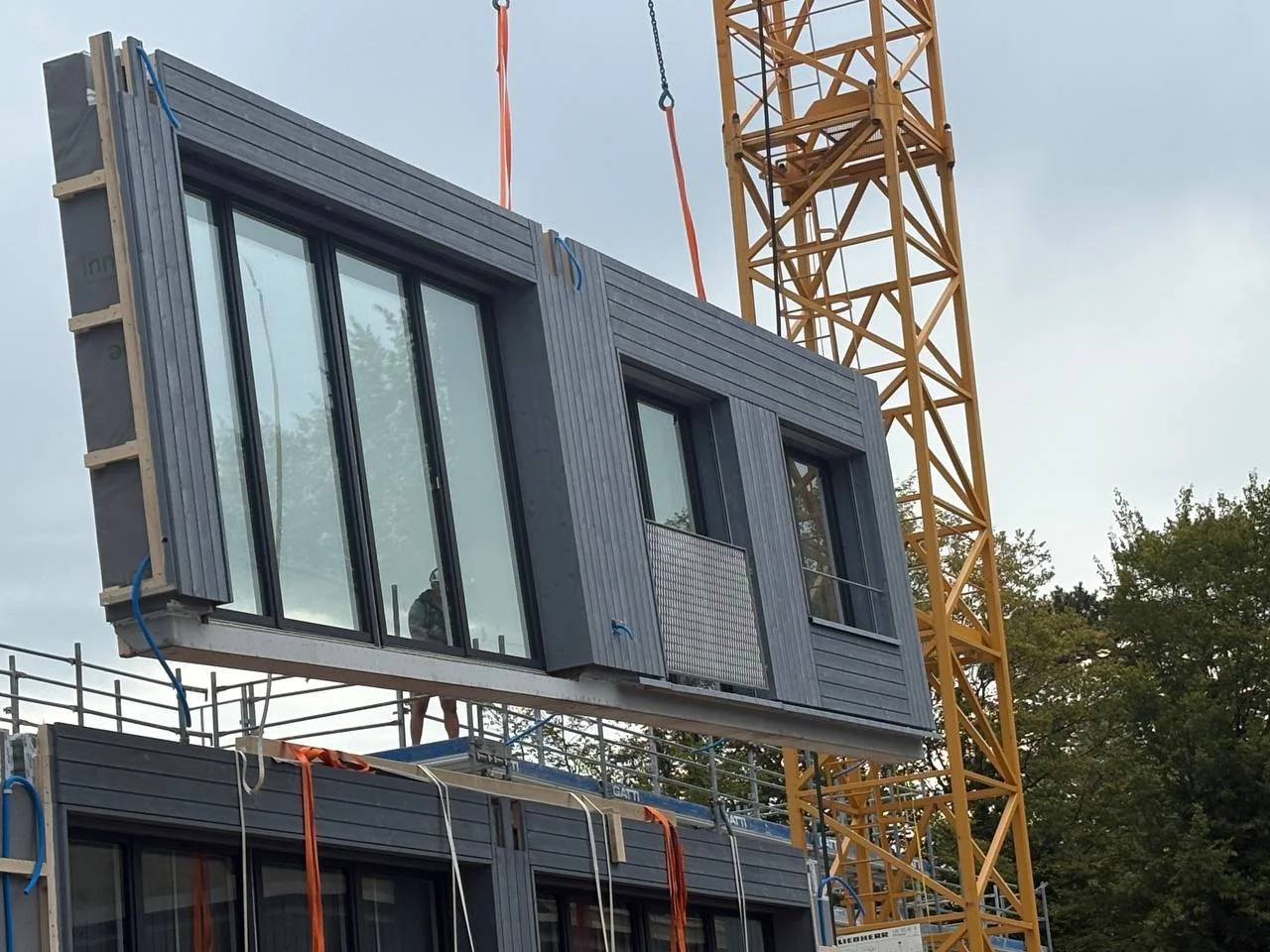 Bau eines modularen Gebäudes mit Holzfassade und großen Fenstern auf einer Baustelle, mit einem gelben Kran im Hintergrund.