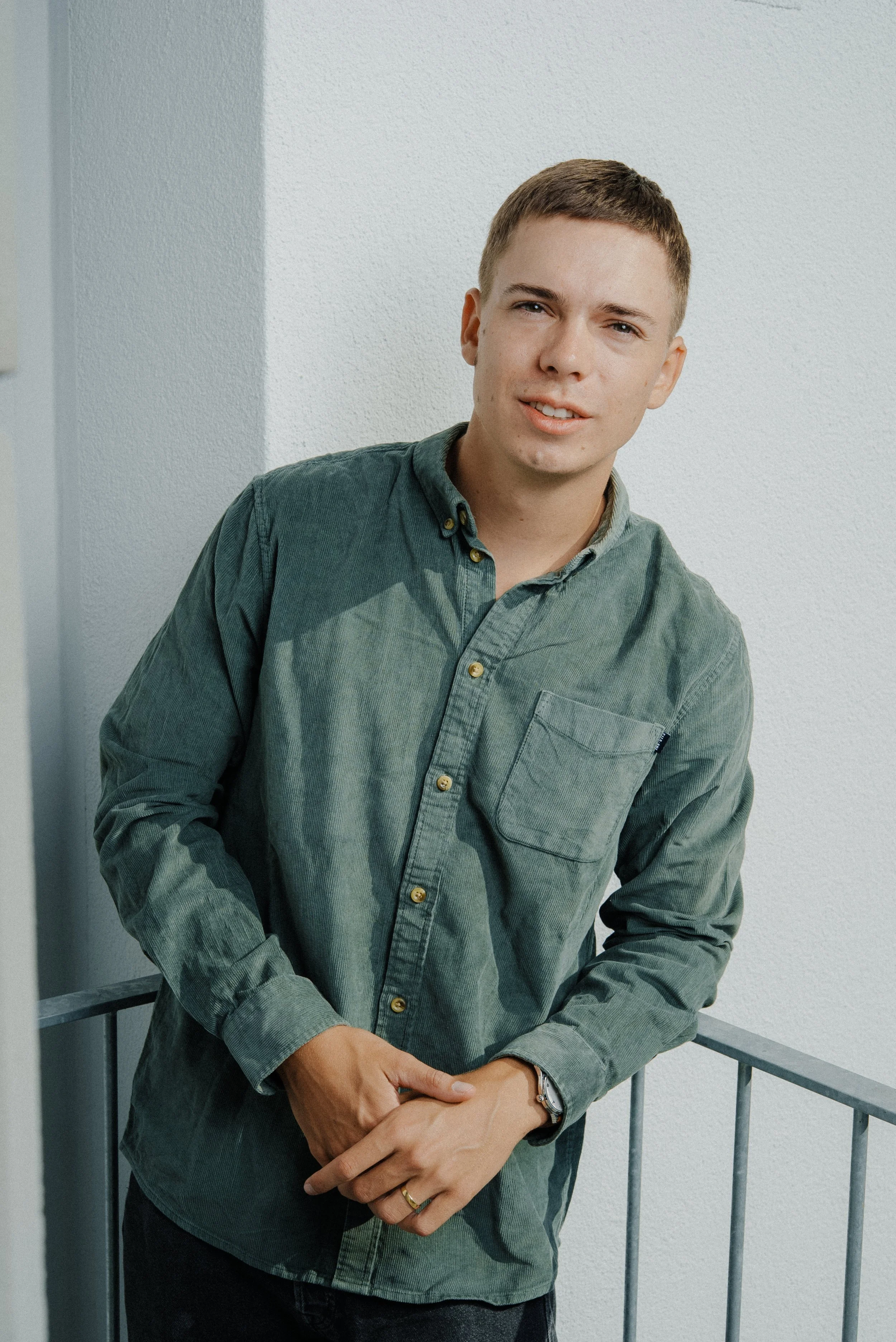 Junge Mann steht an einer Metallgeländer, trägt eine grüne Hemd und eine Armbanduhr, vor einer weißen Wand.
