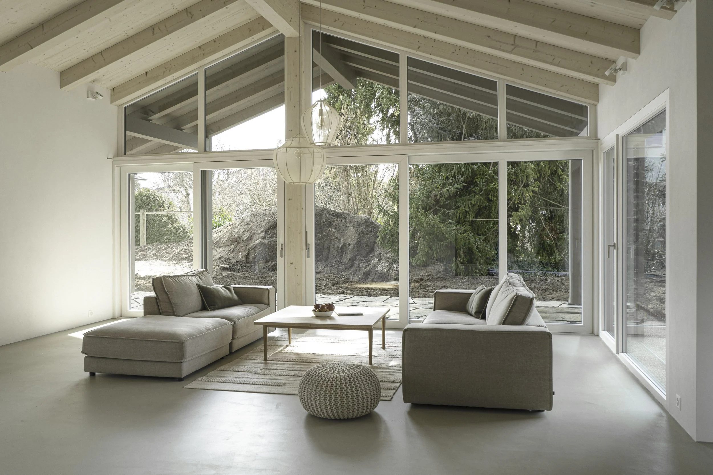 Heller offener Wohnraum mit großen Fenstern, moderner Sofa, einem kleinen Tisch und einem dekorativen Sitzkissen, Blick nach draußen auf eine Gartenlandschaft mit Bäumen.
