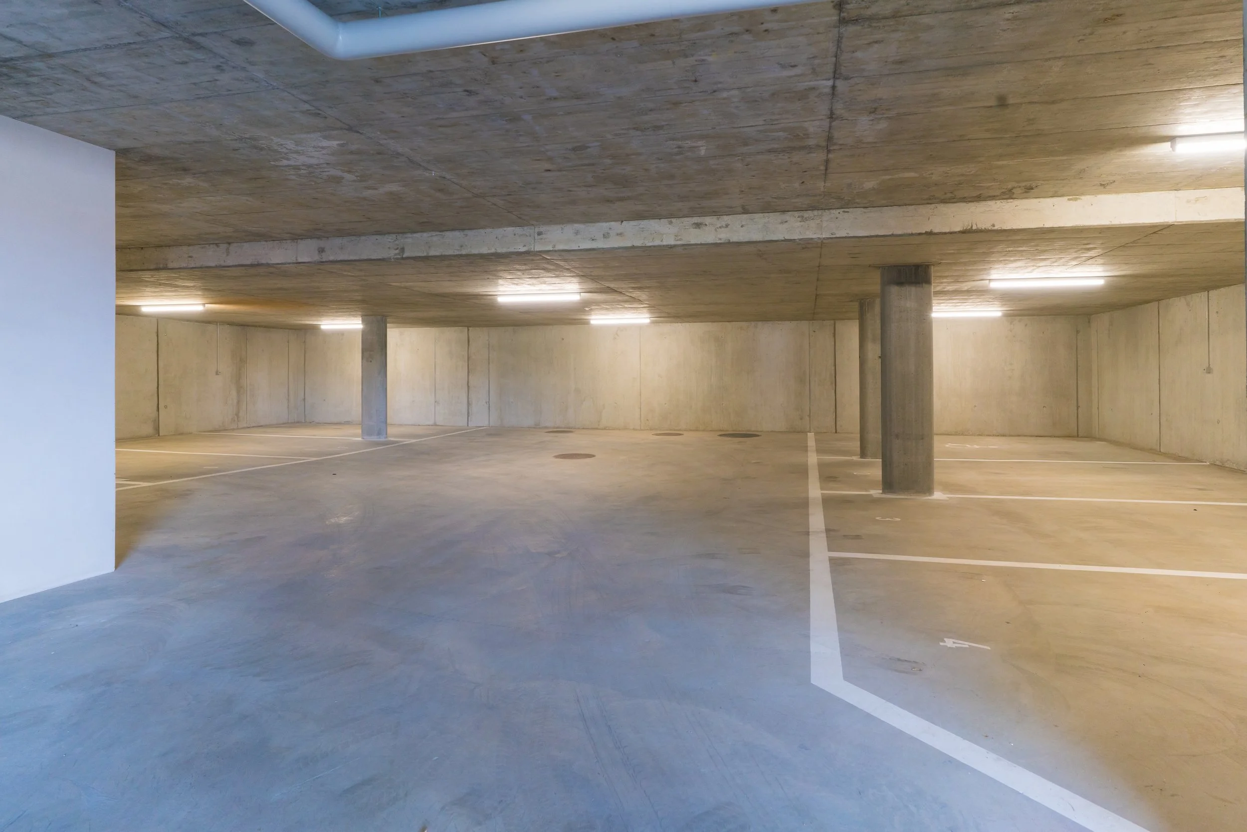Ein leeres, unterirdisches Parkhaus mit Betonwänden und -decken, Parkplätzen, Beleuchtung und Stützpylonen.