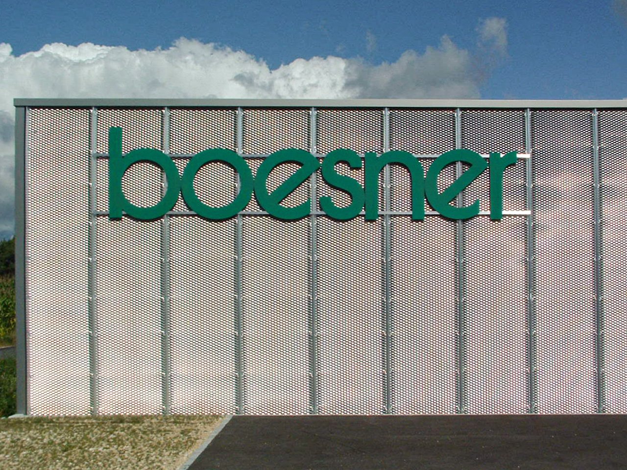 Außenansicht eines Gebäudes mit dem grünen Schriftzug 'boesner' auf einer perforierten Metallfassade, im Hintergrund blauer Himmel mit Wolken.