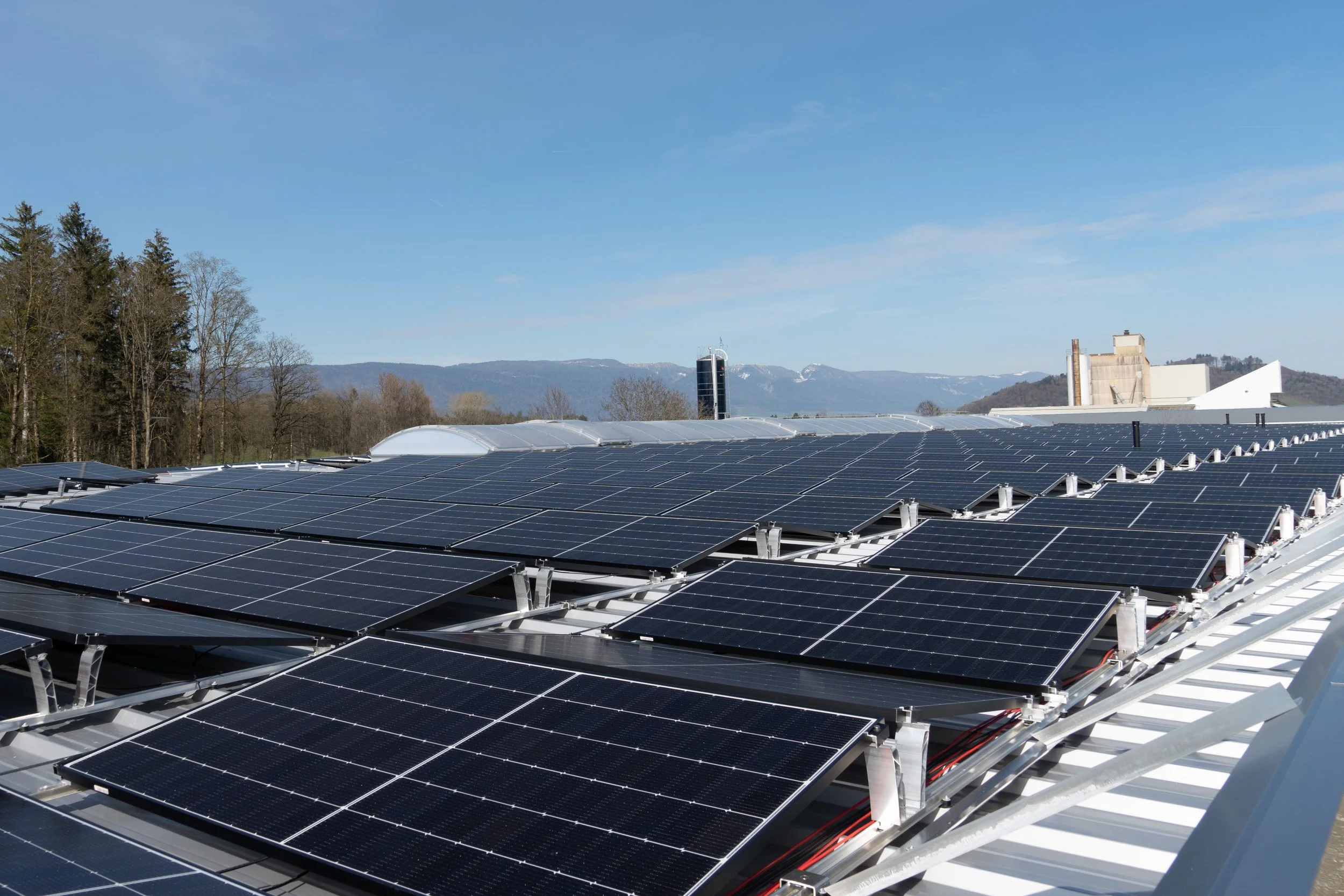 Photovoltaikanlagen auf einem Dach mit Wald und Bergen im Hintergrund bei strahlendem Himmel.